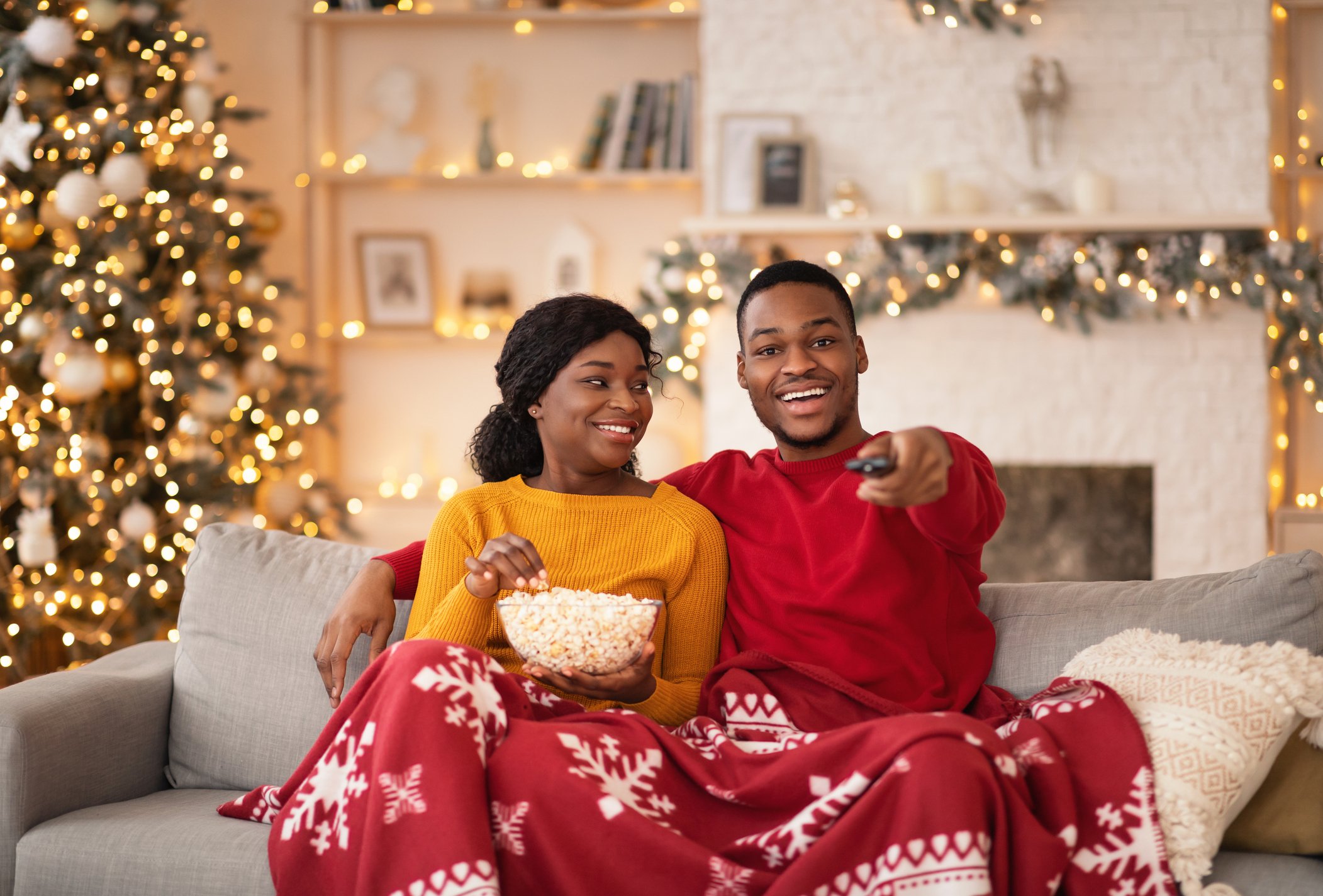 Tv programs and films at new years night at home. Young smiling african american guy switches channels with remote control, lady holds popcorn, couple covered with blanket in interior with Xmas tree