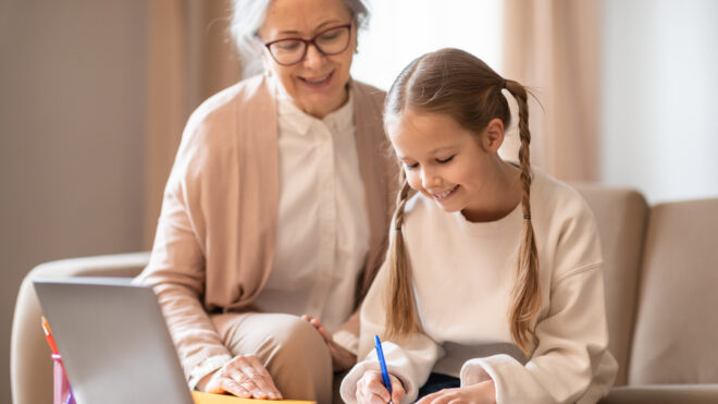 A senior woman with glasses is helping a young girl, who is focused on writing in her notebook, with her homework at a table. They are using a laptop
