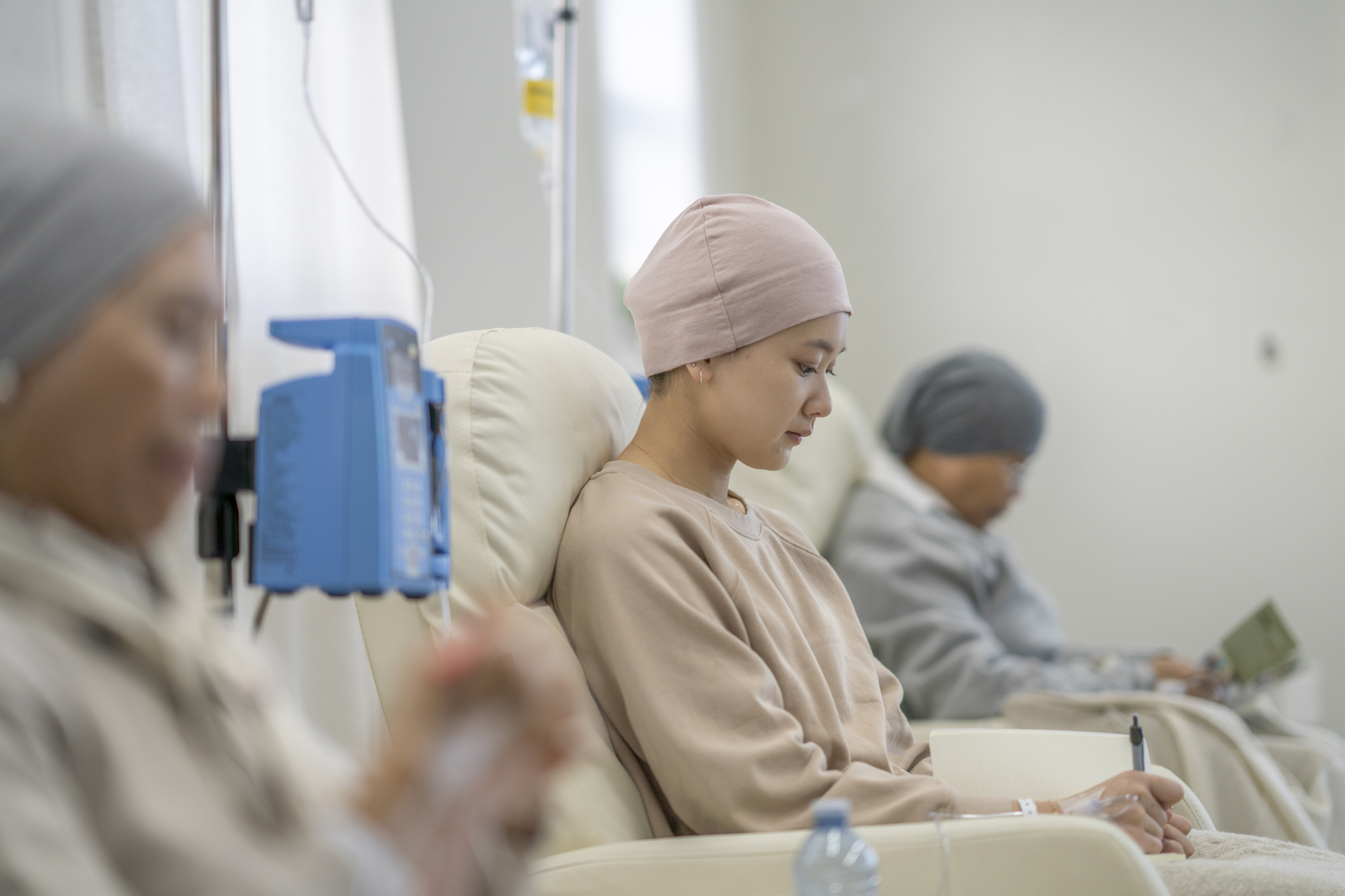 Three women sit in individual chairs as they receive their Chemotherapy by intravenous. They are each dressed comfortably and have head scarves on to keep them warm as they engage in the activity they each brought to keep them occupied.