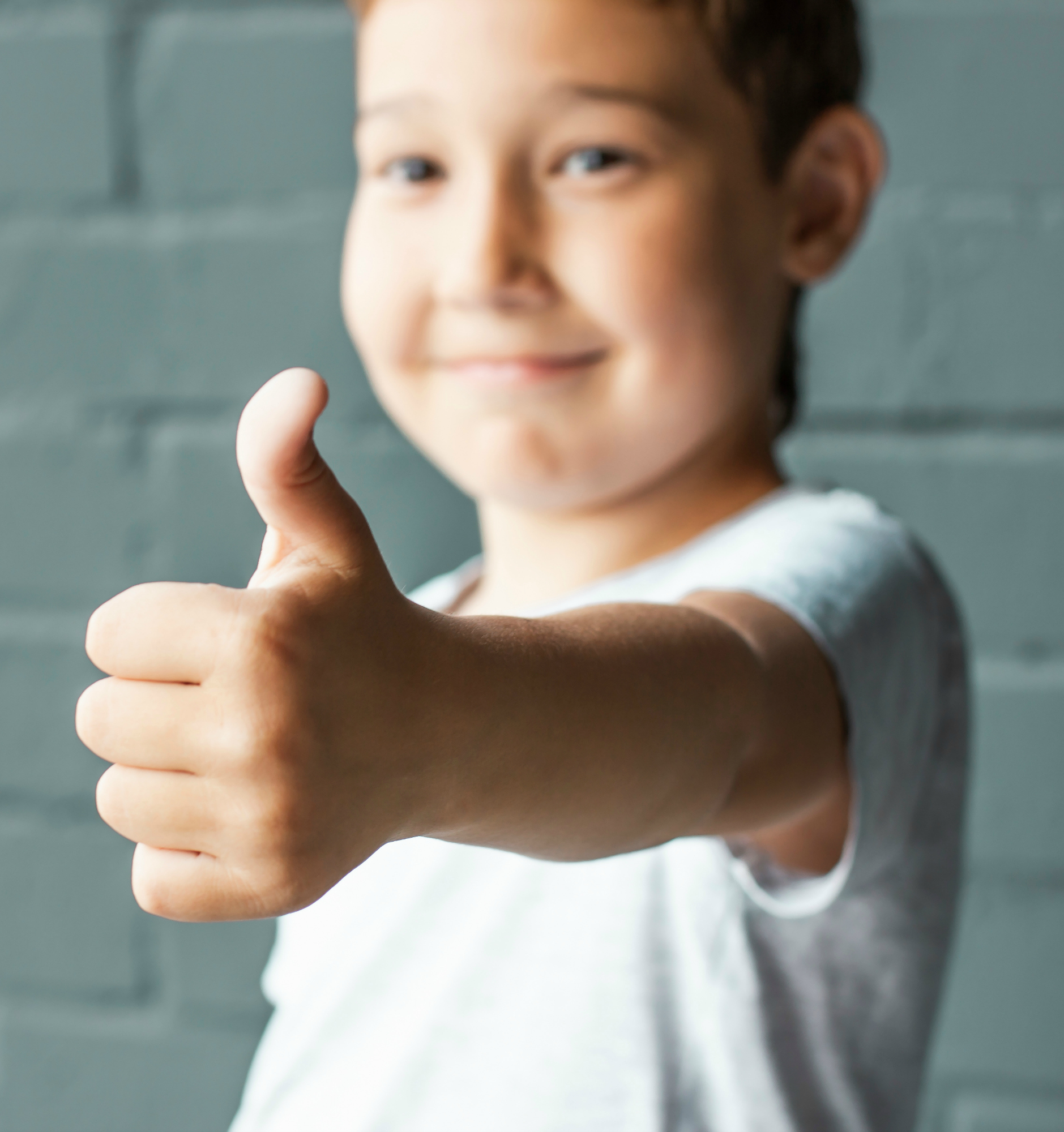 Cute smiling boy 8 years old showing thumbs up on background of grey brick wall