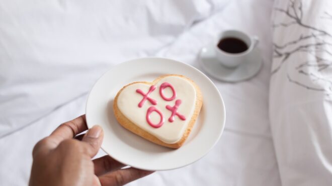 hand-holding-a-plate-with-a-heart-shaped-valentines-day-cookie-with-xoxo-lettering-in-bed_t20_mvkzxe
