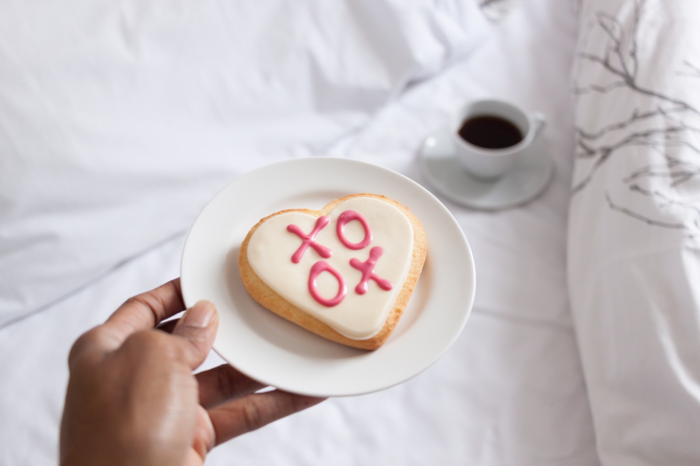 hand-holding-a-plate-with-a-heart-shaped-valentines-day-cookie-with-xoxo-lettering-in-bed_t20_mvkzxe