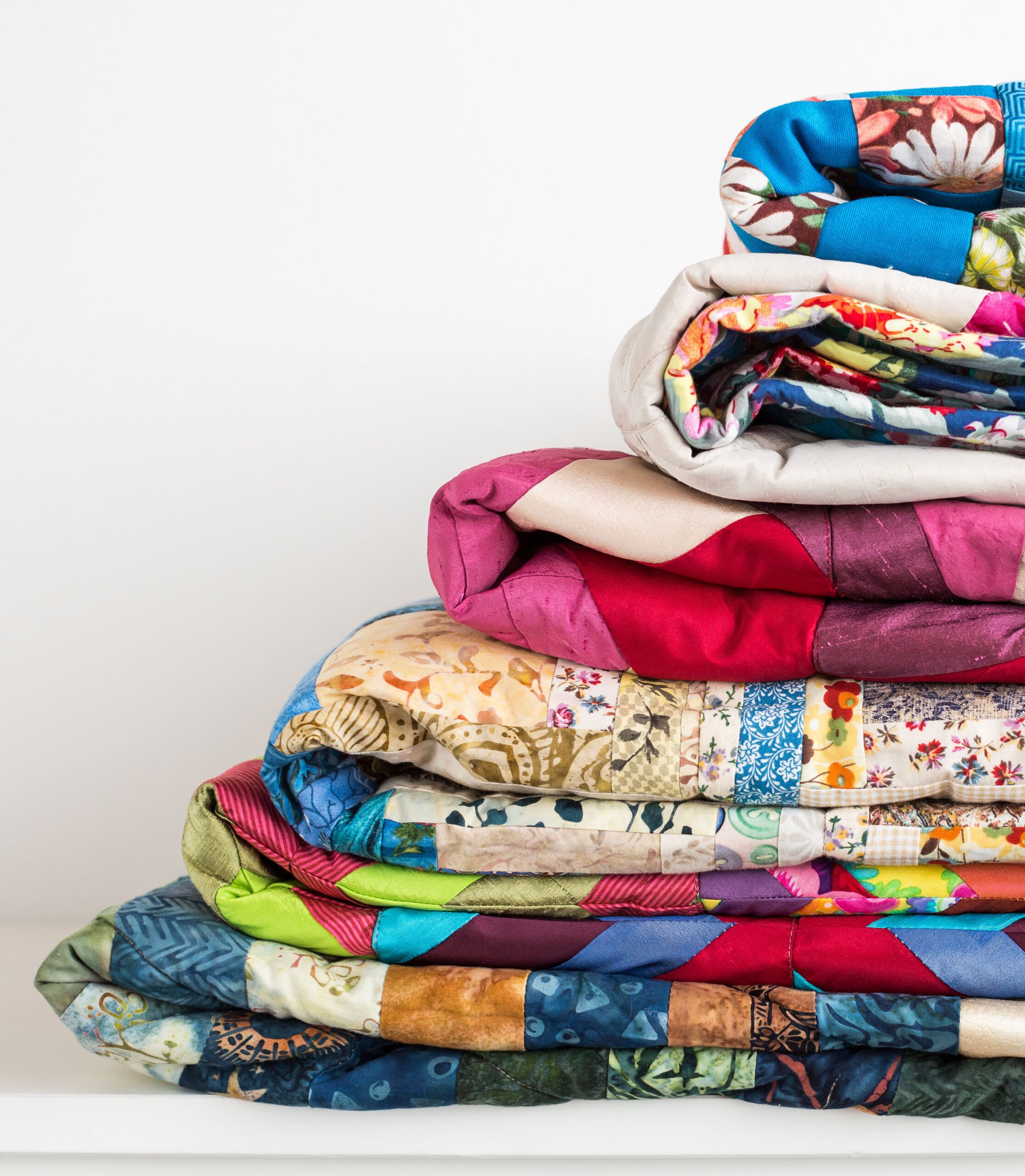 sewing, patchwork and fashion concept - beautiful colorful quilts were neatly folded and stored in several rows in height for storage, sale of finished textile stitched products on white background
