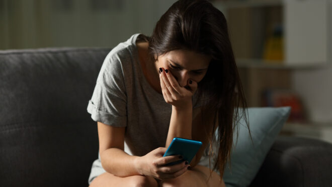 shocked-woman-couch-phone - Sad teen receiving bad news online in a smart phone sitting on a couch in the living room at home