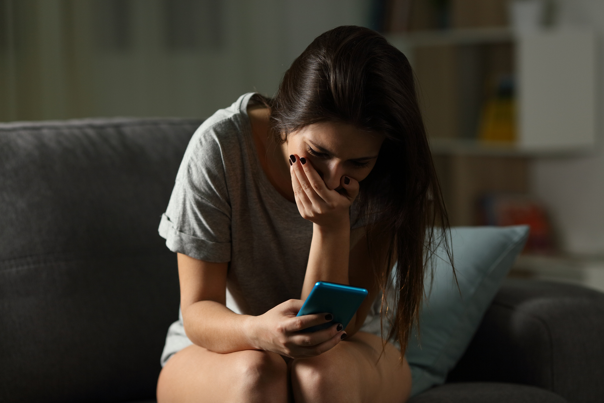 shocked-woman-couch-phone - Sad teen receiving bad news online in a smart phone sitting on a couch in the living room at home