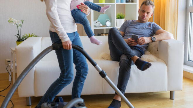 Woman with small child doing housekeeping while man sitting in couch - Woman with small child in her hands doing housekeeping while man sitting in couch and relaxing.