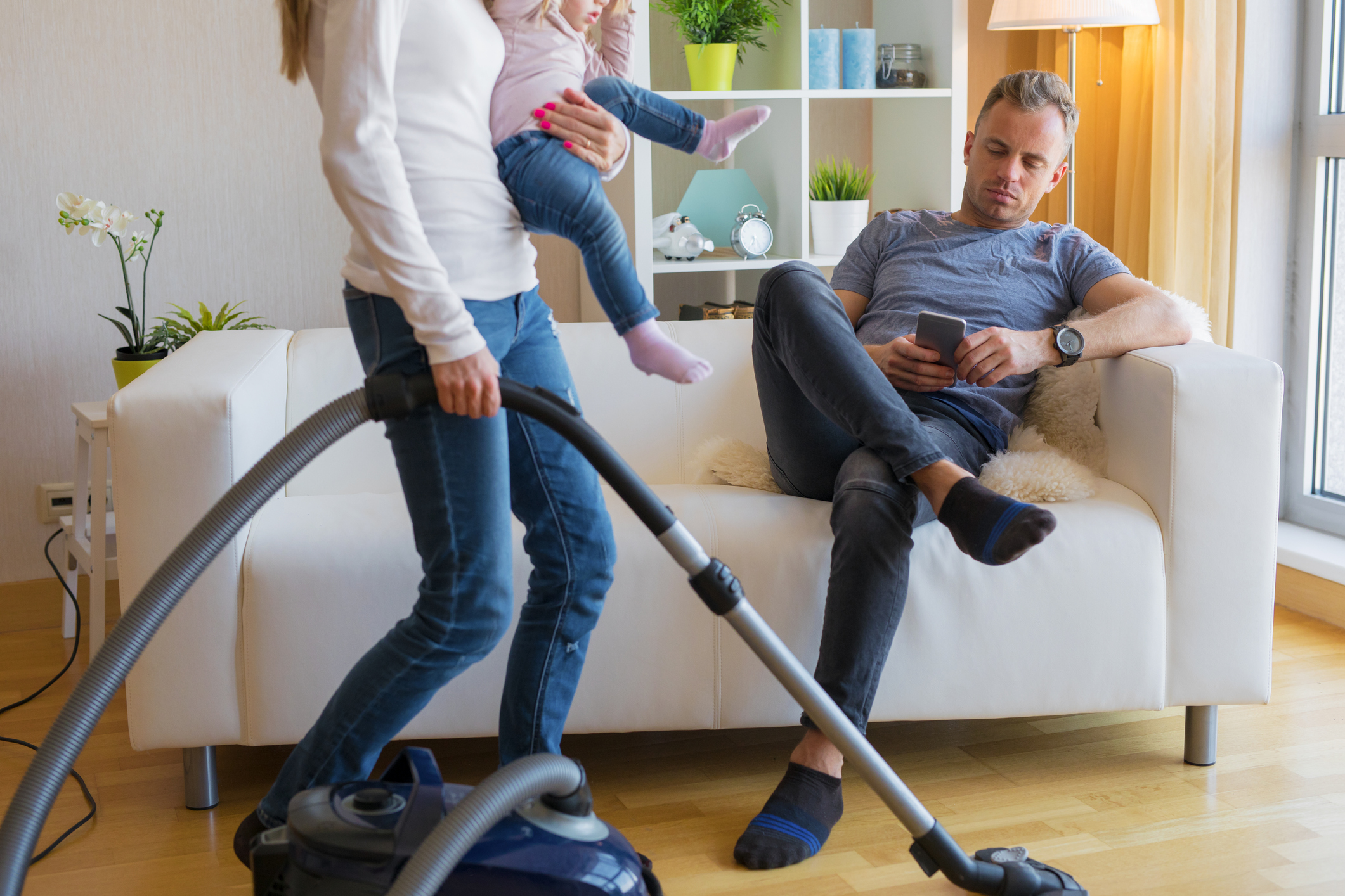 Woman with small child doing housekeeping while man sitting in couch - Woman with small child in her hands doing housekeeping while man sitting in couch and relaxing.