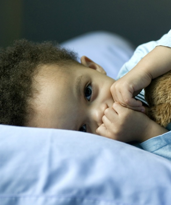 Baby boy lying in bed with teddy bear, hands over mouth, looking at camera