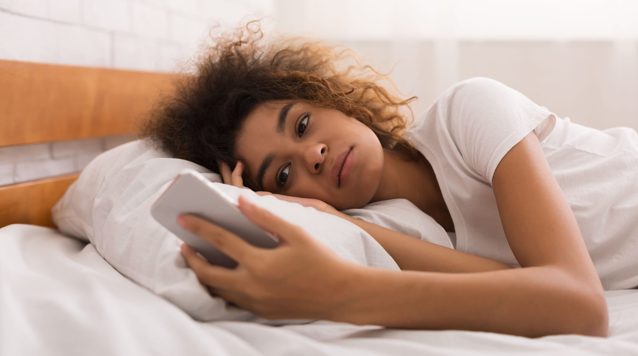 Woman looking unhappy at text message, lying in bed
