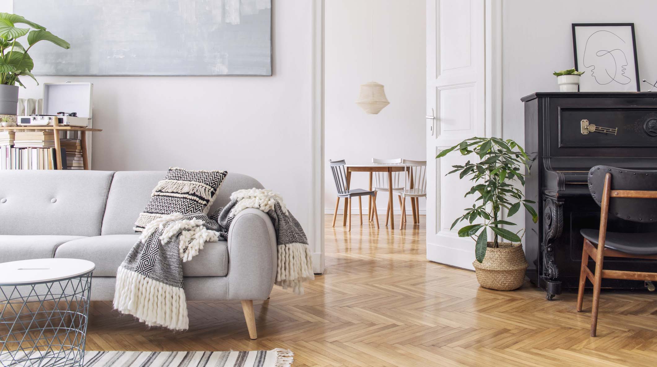 Modern scandianvian decor of living room with design sofa with elegant blanket, coffee table and bookstand on the white wall. Brown wooden parquet. Concept of minimalistic interior with piano. Mock up