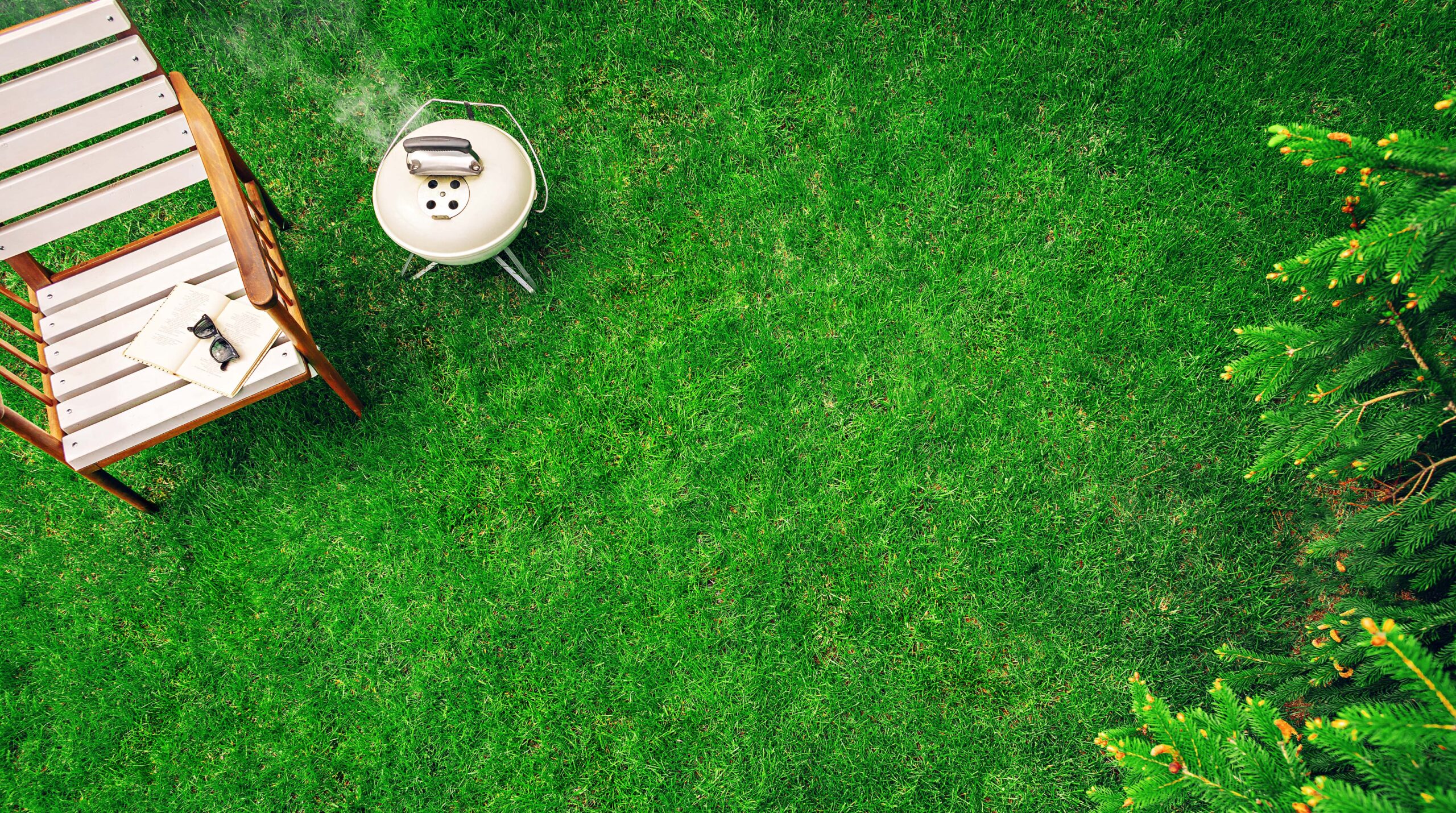 Ivory colored grill on the grass near the wooden armchair with a book and glasses. Top view