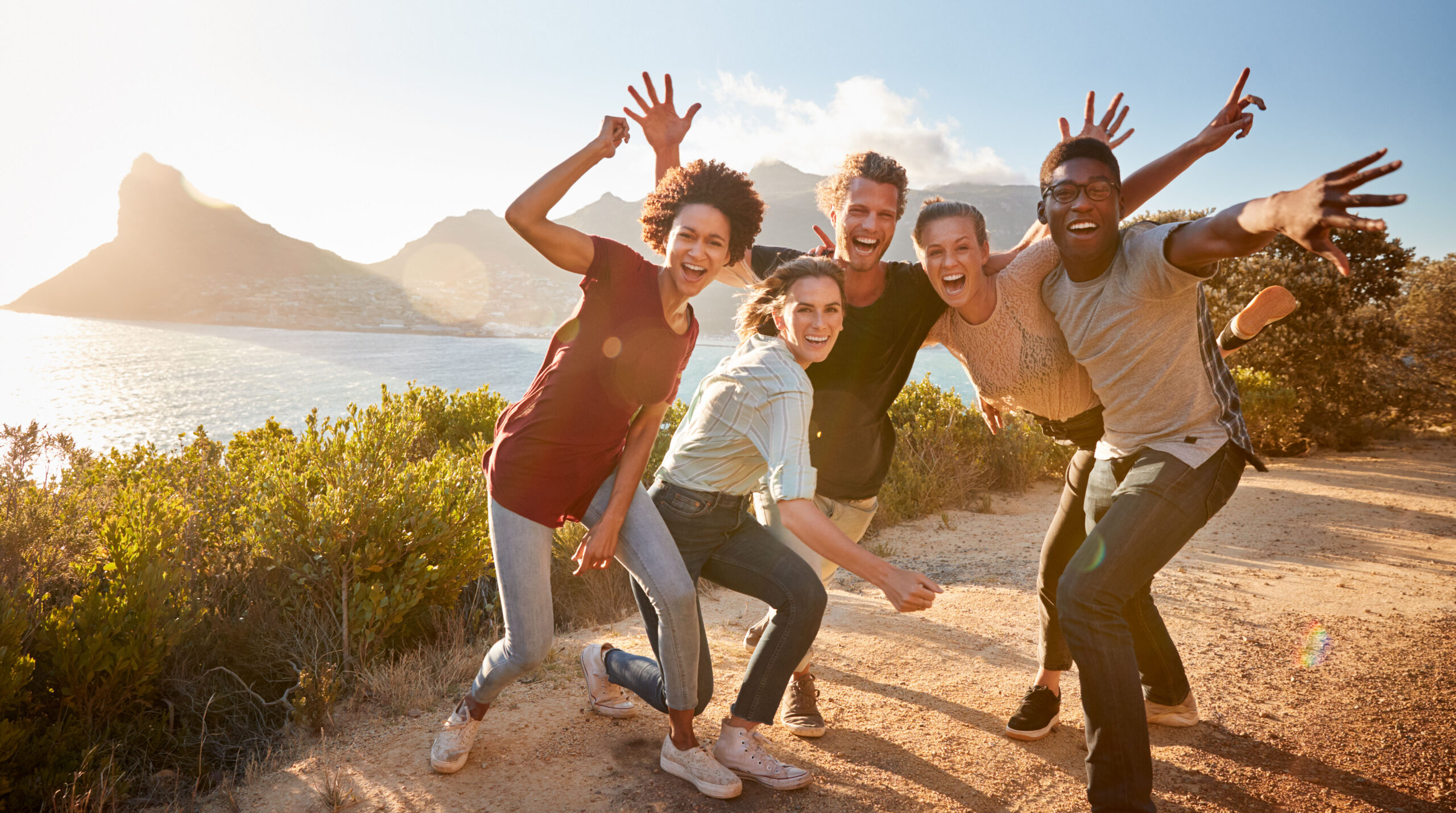 Five millennial friends on a road trip have fun posing for photos on a coastal path, full length
