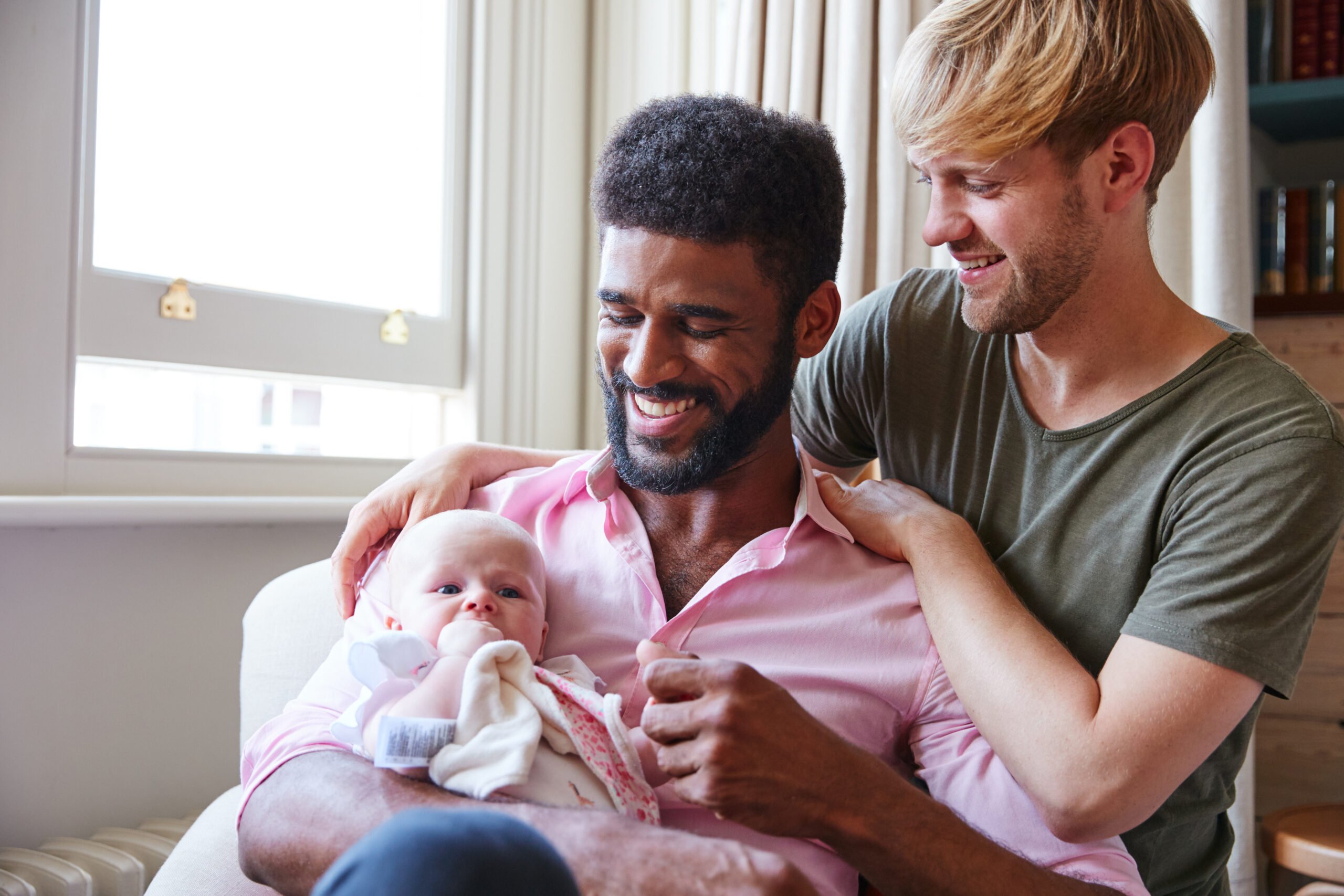 Loving Male Same Sex Couple Cuddling Baby Daughter On Sofa At Home Together