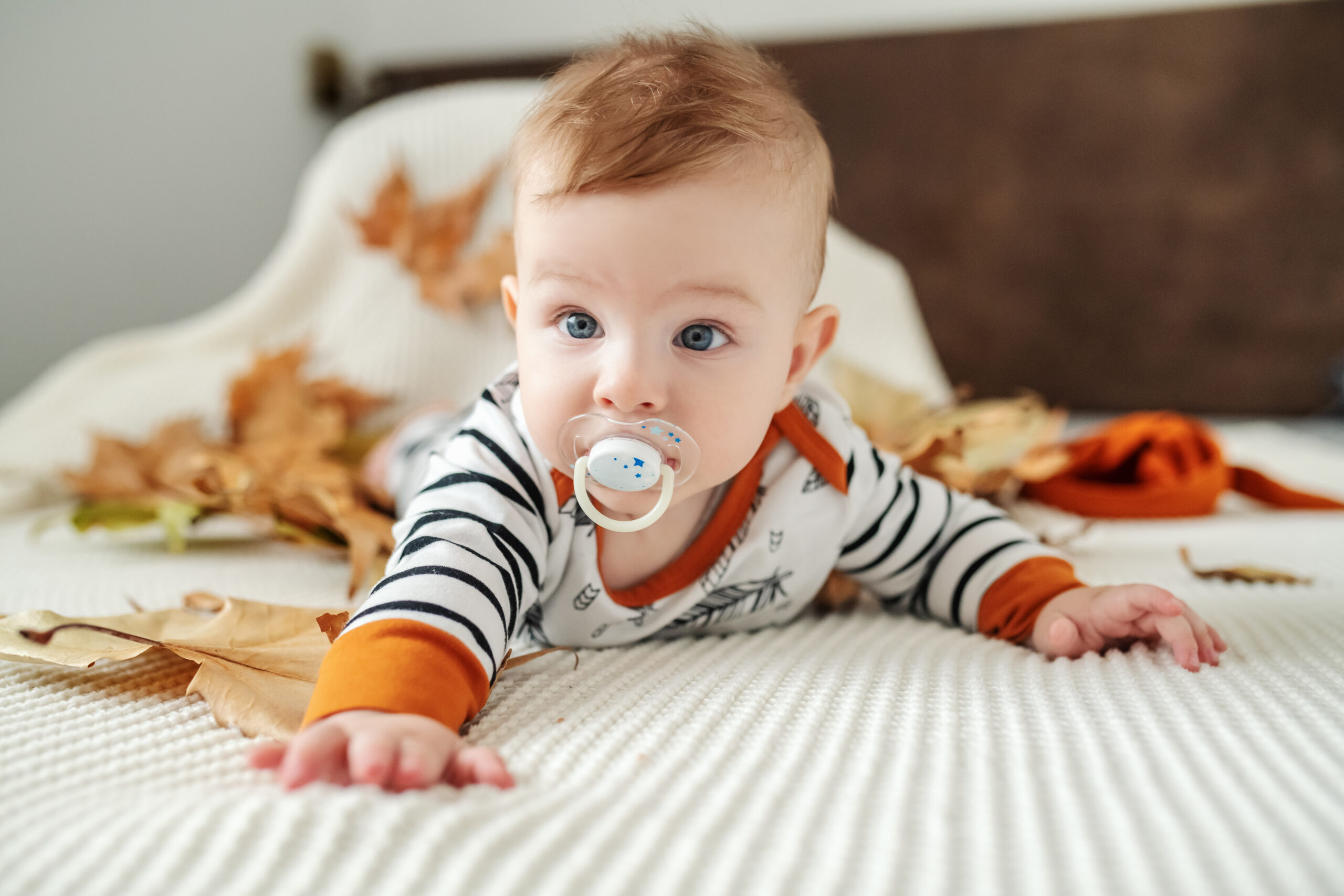 Cute baby boy with pacifier lying down on stomach on bed surrounded by autumn leaves.