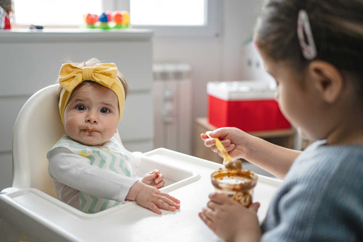 Cute baby sitting in a high chair being fed by her sister