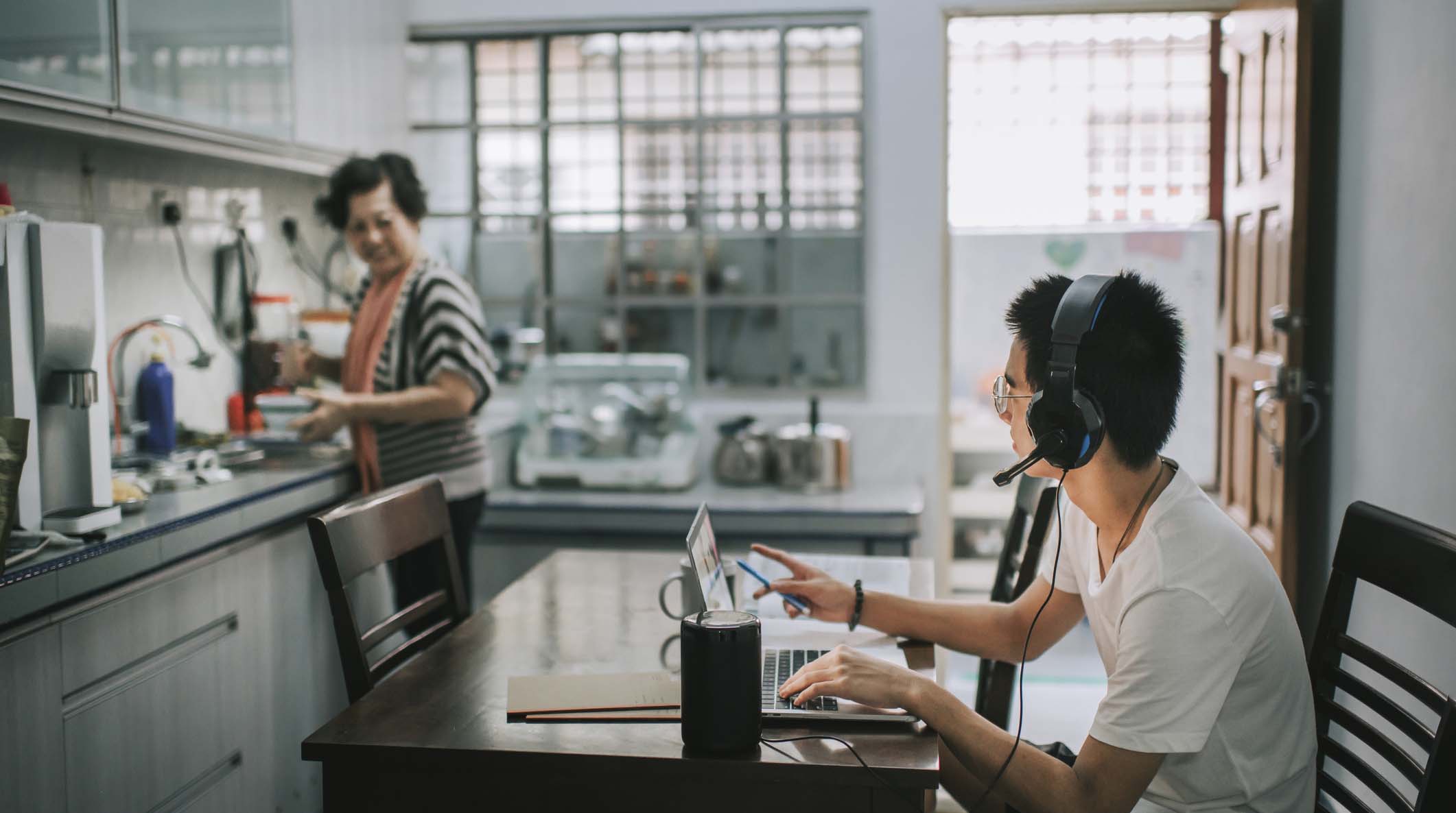 An asian chinese teenager chatting online with all his classmates via online software while doing his homework at the kitchen dining table while his grandmother cooking in the kitchen preparing food