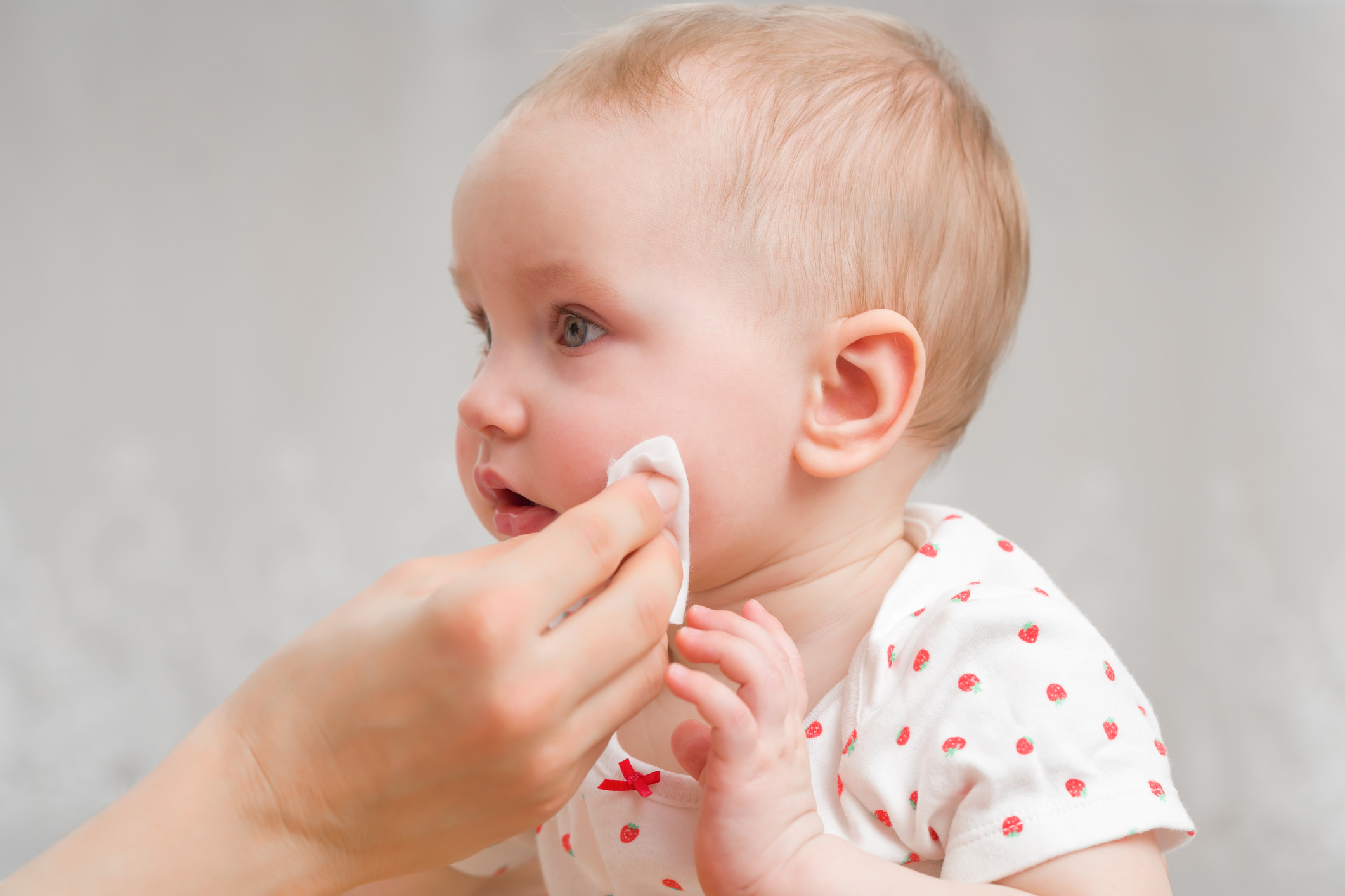 Young mother hand wiping baby girl cheek with white cotton pad. Daily routine. Care about infant clean and soft body skin. Closeup. Side view.
