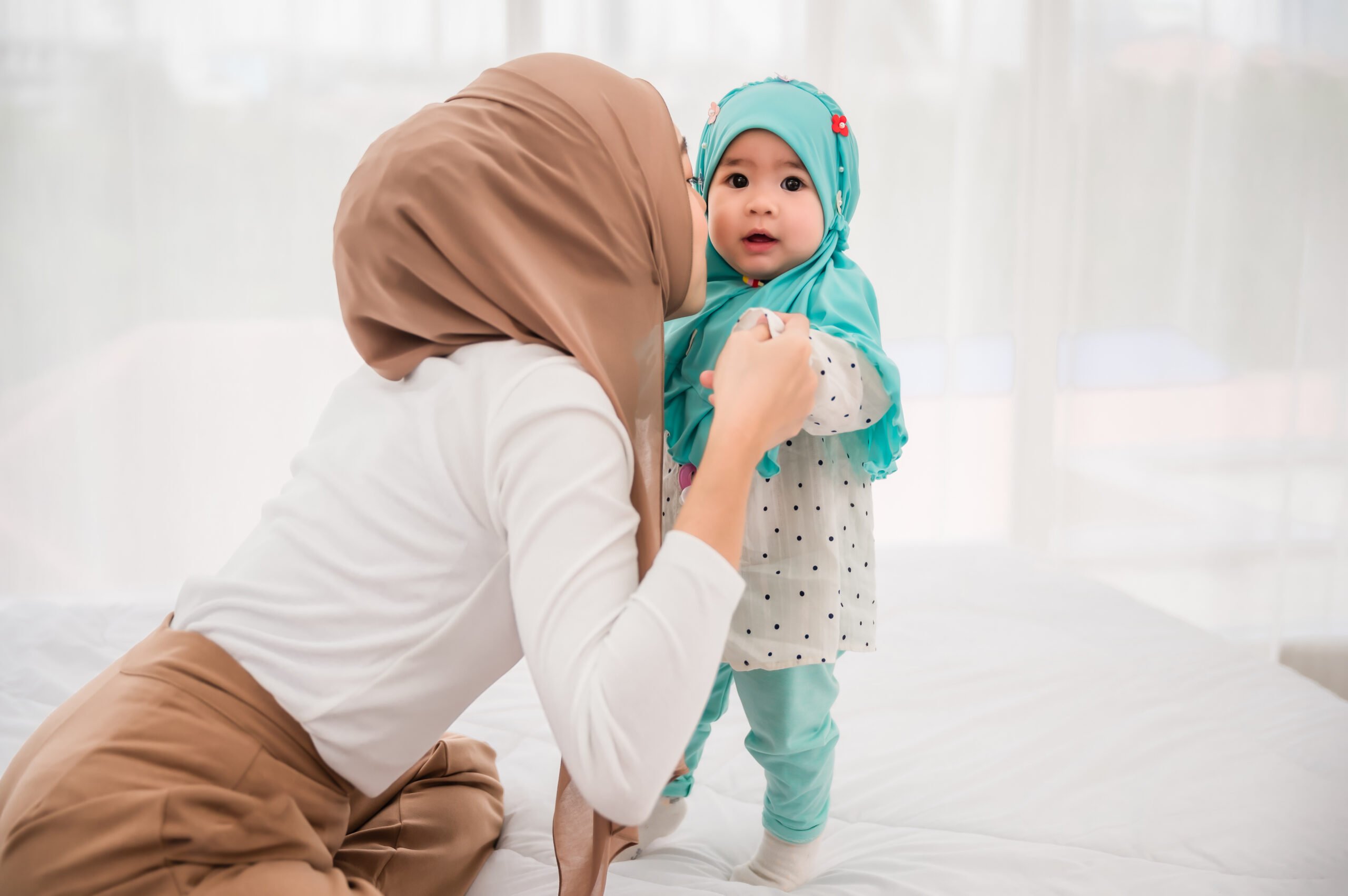 Happy muslim mother and adorable little baby daughter in hijab on bed in white bedroom at home. Muslim mom hands holding baby for practice standing on bed at indoors. Love of mother and baby concept.