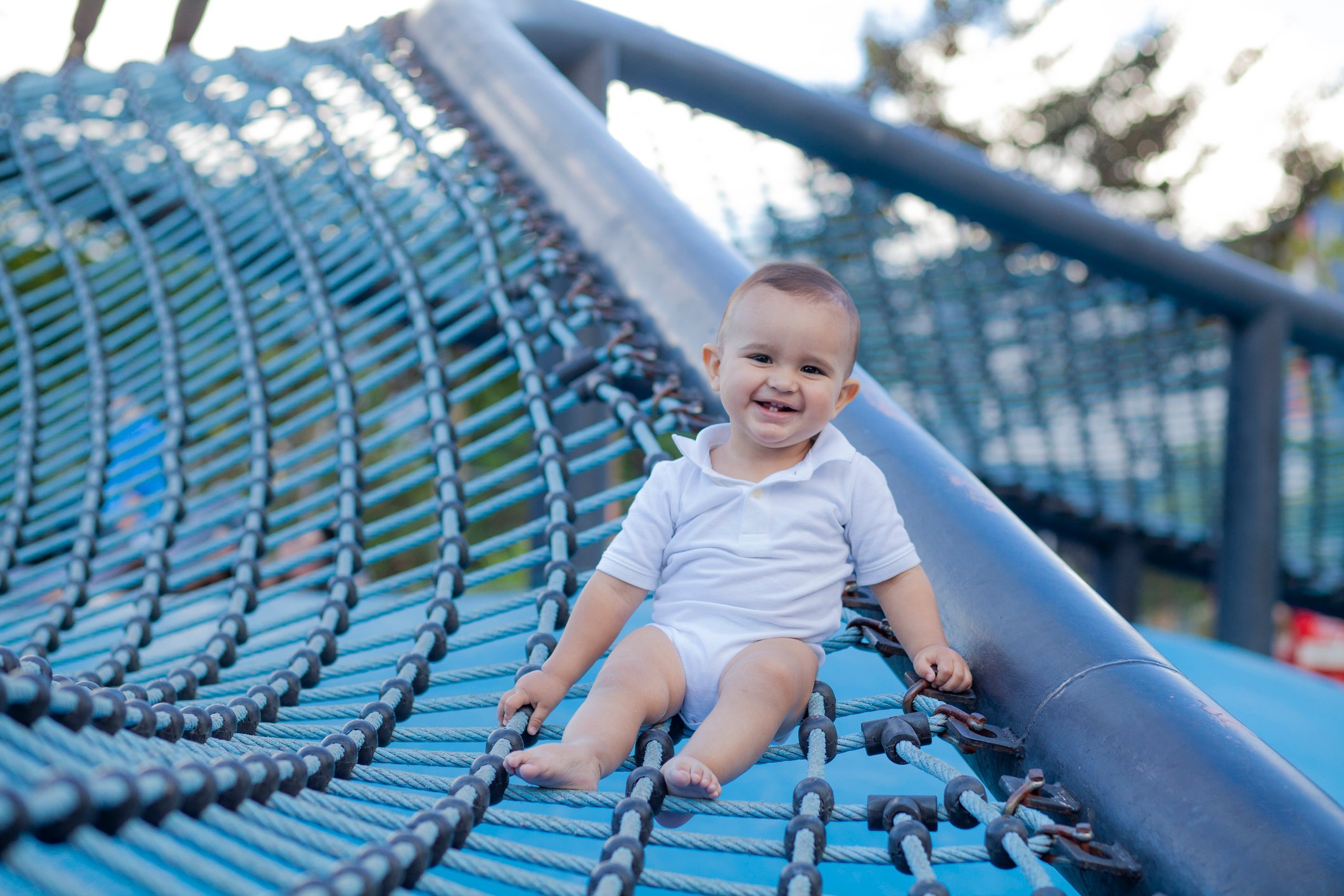 1 year old Latino boy sitting on a raised net in a park stares at the camera and shows a big smile