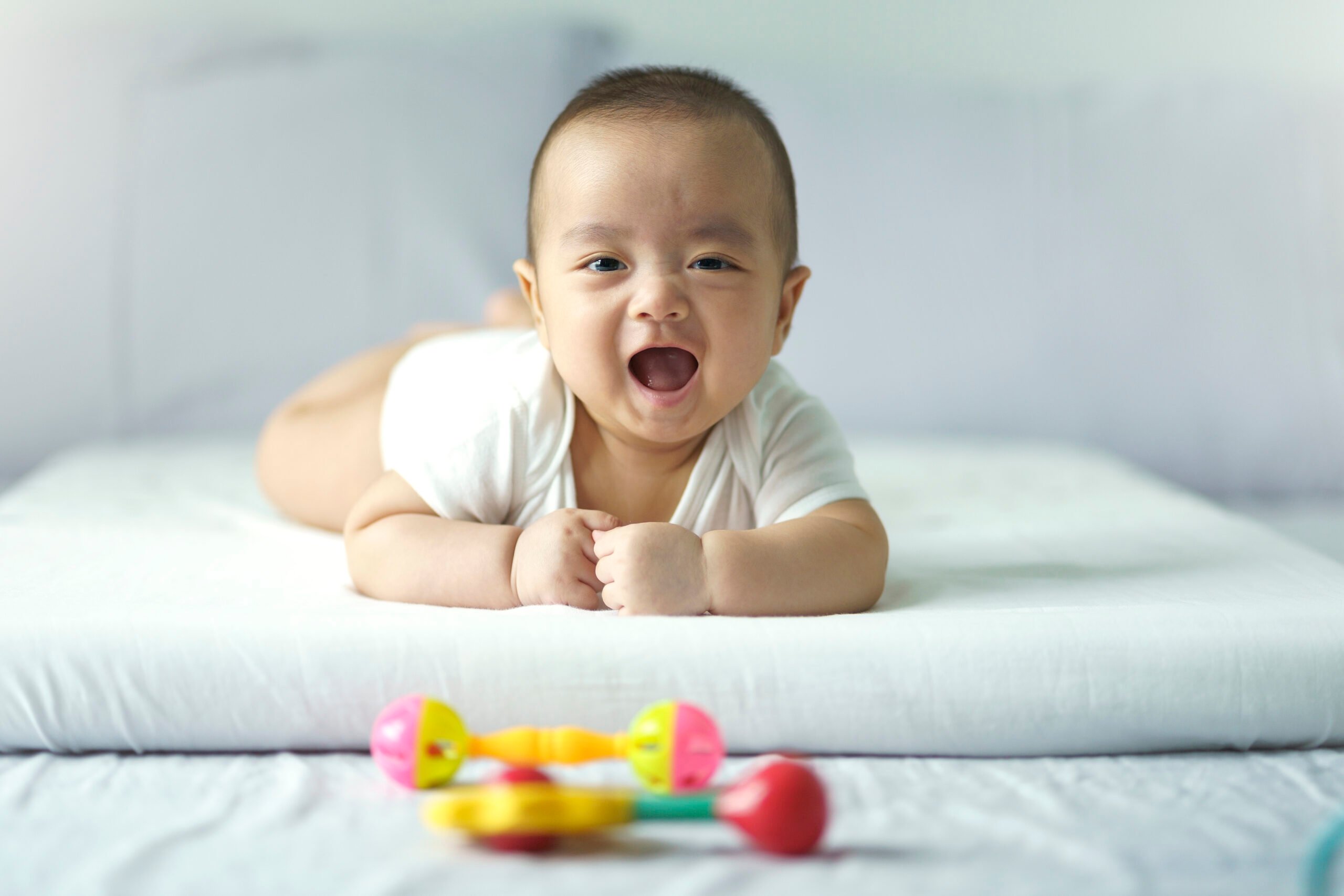 Asian cute baby in white sunny bedroom. Newborn child relaxing on bed. Nursery for young children. Textile and bedding for kid. Family morning at home. New born kid during tummy time with toy.