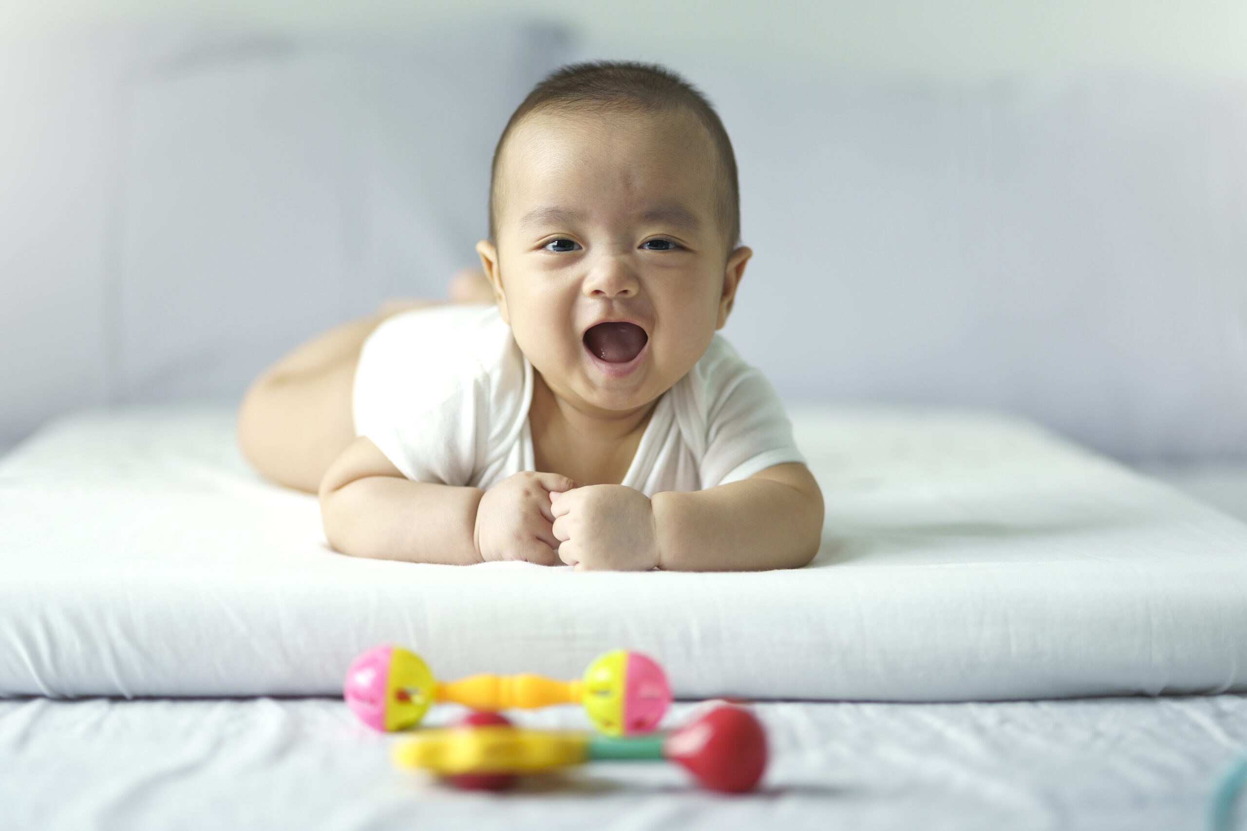 Asian cute baby in white sunny bedroom. Newborn child relaxing on bed. Nursery for young children. Textile and bedding for kid. Family morning at home. New born kid during tummy time with toy.