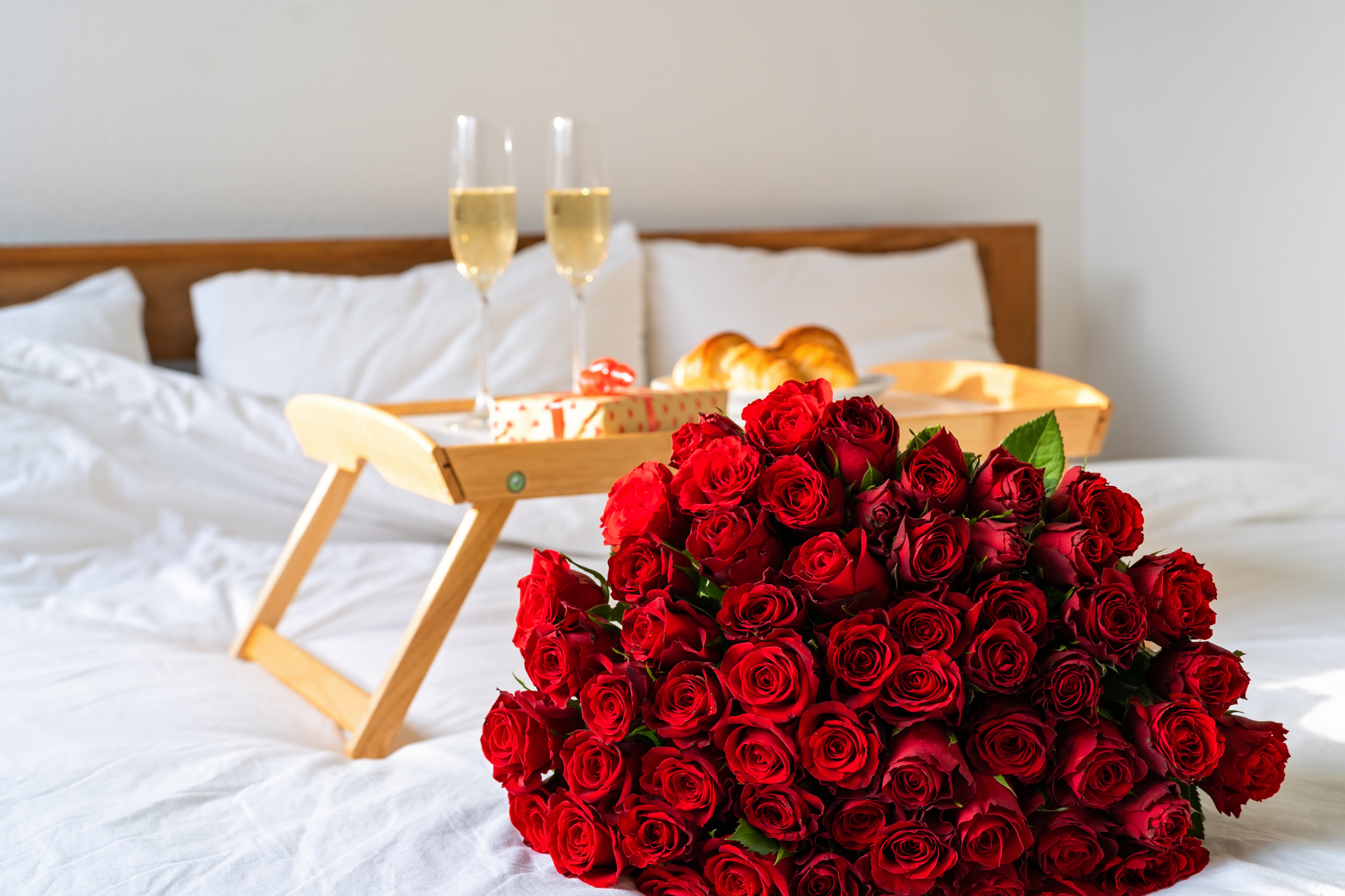 Celebrating Saint Valentine's Day with bouquet of red roses, glass of champagne and croissants on tray on bed. Romantic surprise breakfast for Mother's day or International women day