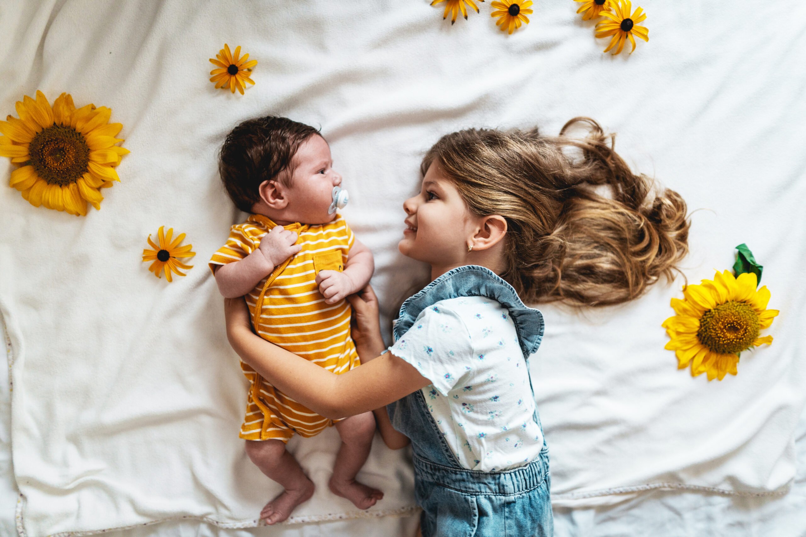 Little girl lying on bed next to her baby brother, surrounded with sunflowers