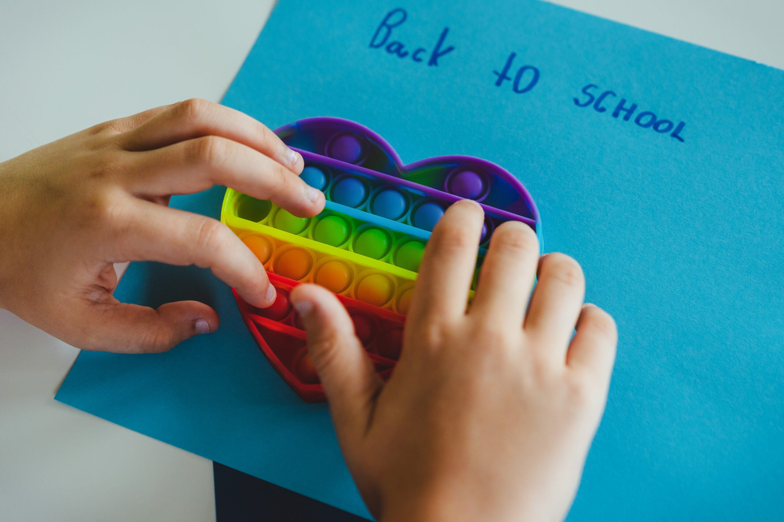 Back to school. Child playing with colourful heart shape push pop it toy. Anti-stress sensory game and text "Back to school" written kid's hand on blue background. Reduce school-related anxious, stress.