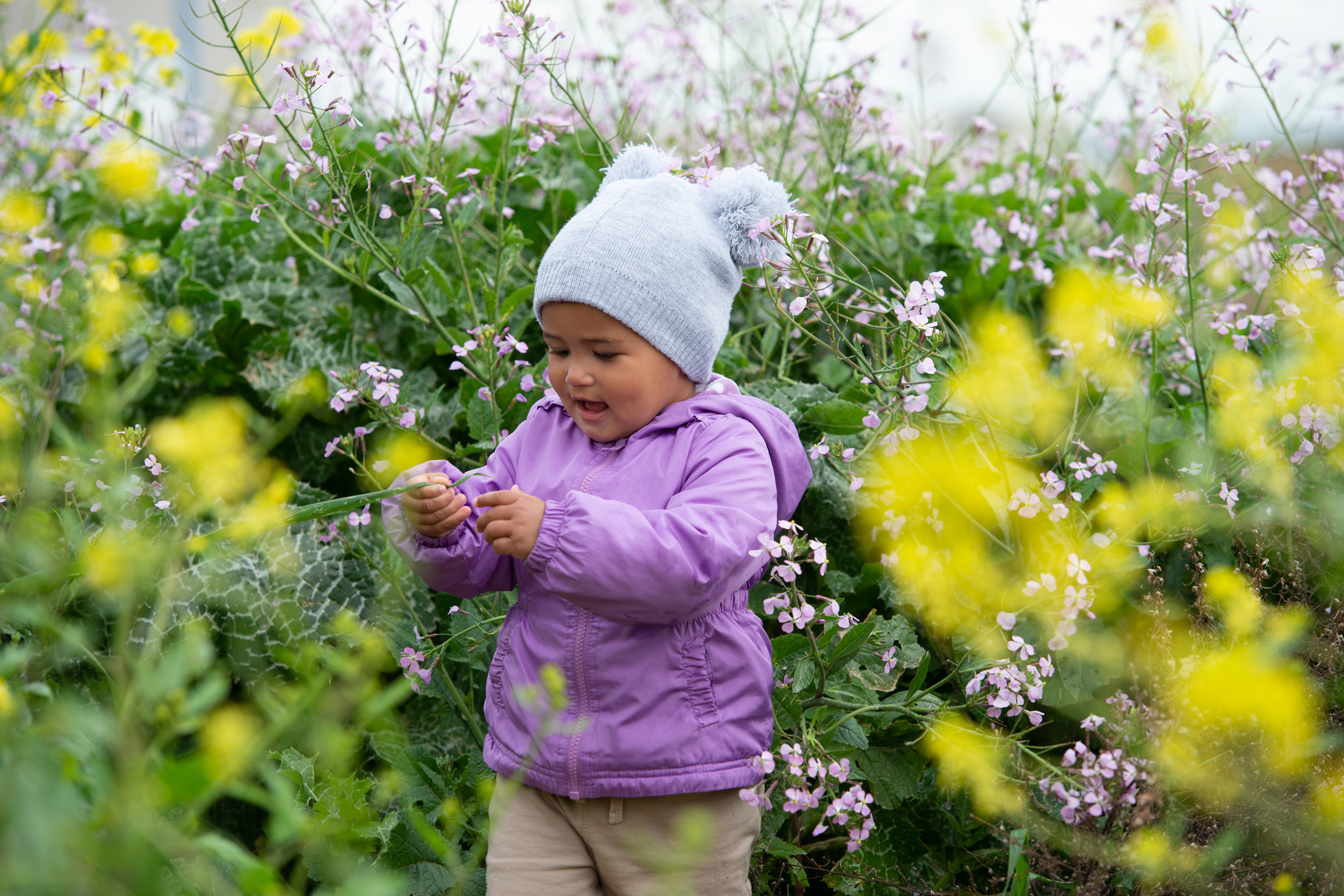 Little curious baby girl among blooming flowers plants.