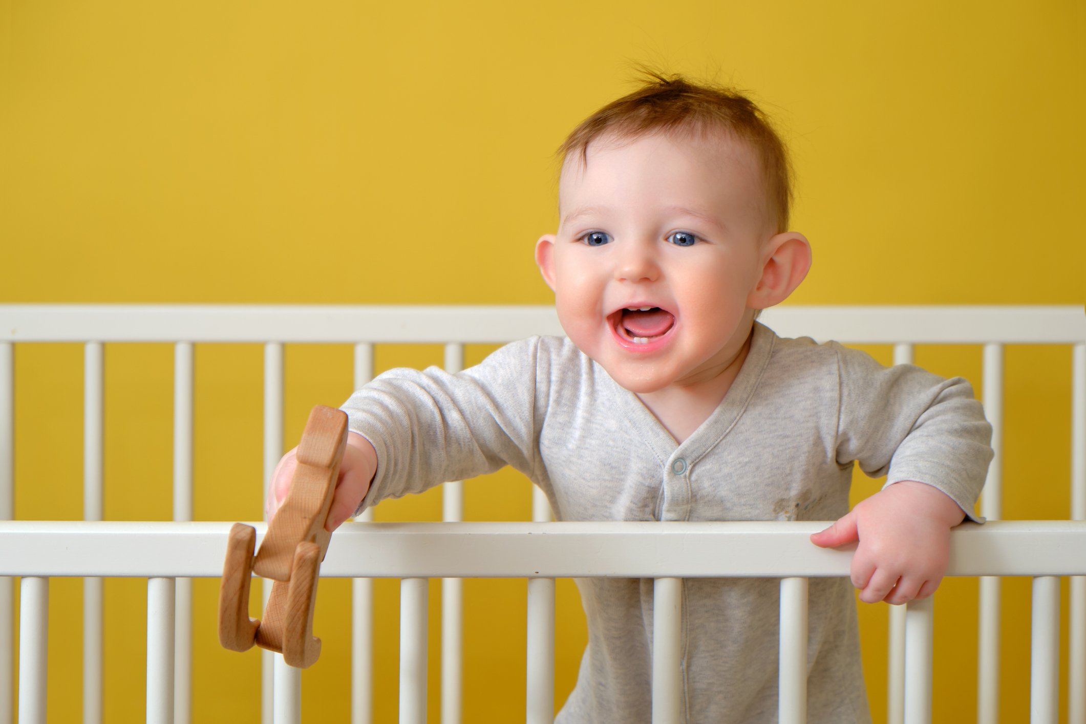 Happy toddler baby boy in a crib with a wooden toy in his hand, yellow studio background. A smiling child in pajamas