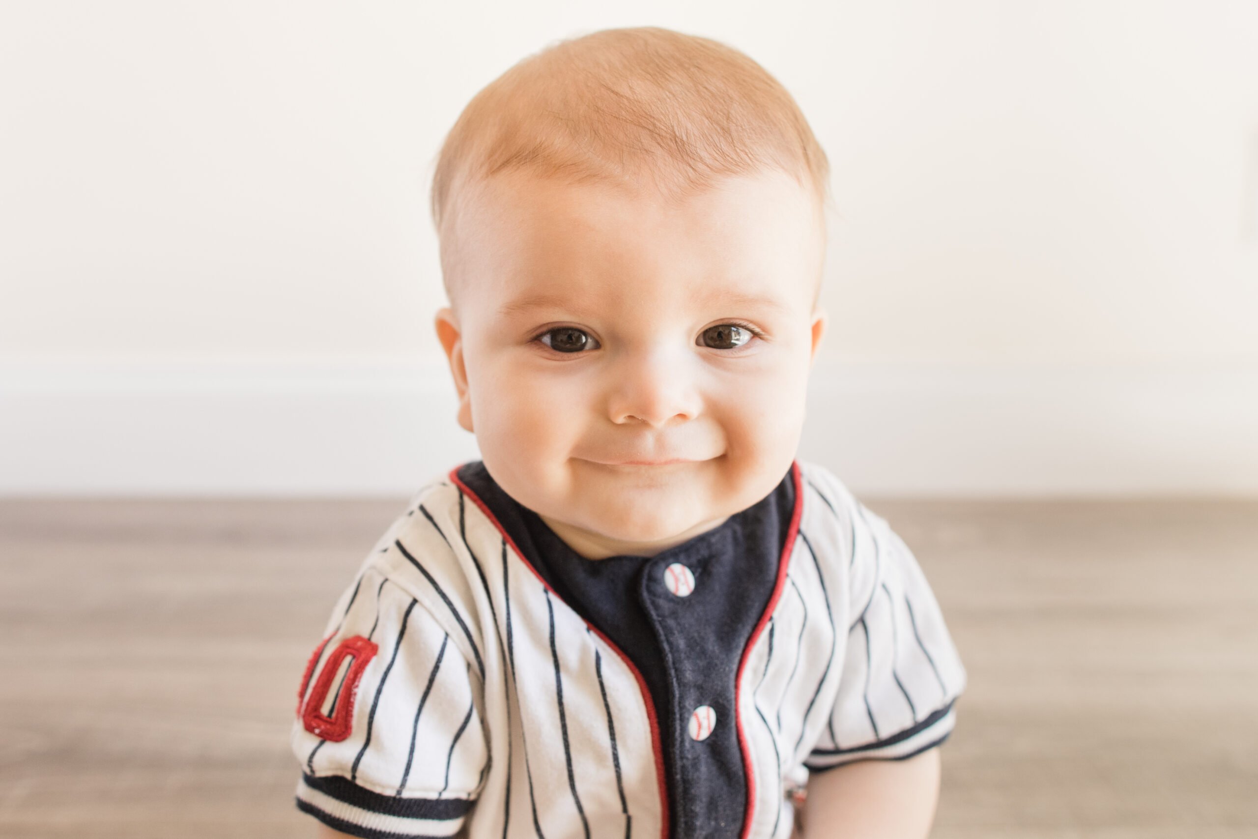 7-Month-Old Baby Boy Dressed in a Baseball Outfit & Playing with a Softball While Learning a New Developmental Skill of Sitting Up on His Own on a Wooden Floor at Home