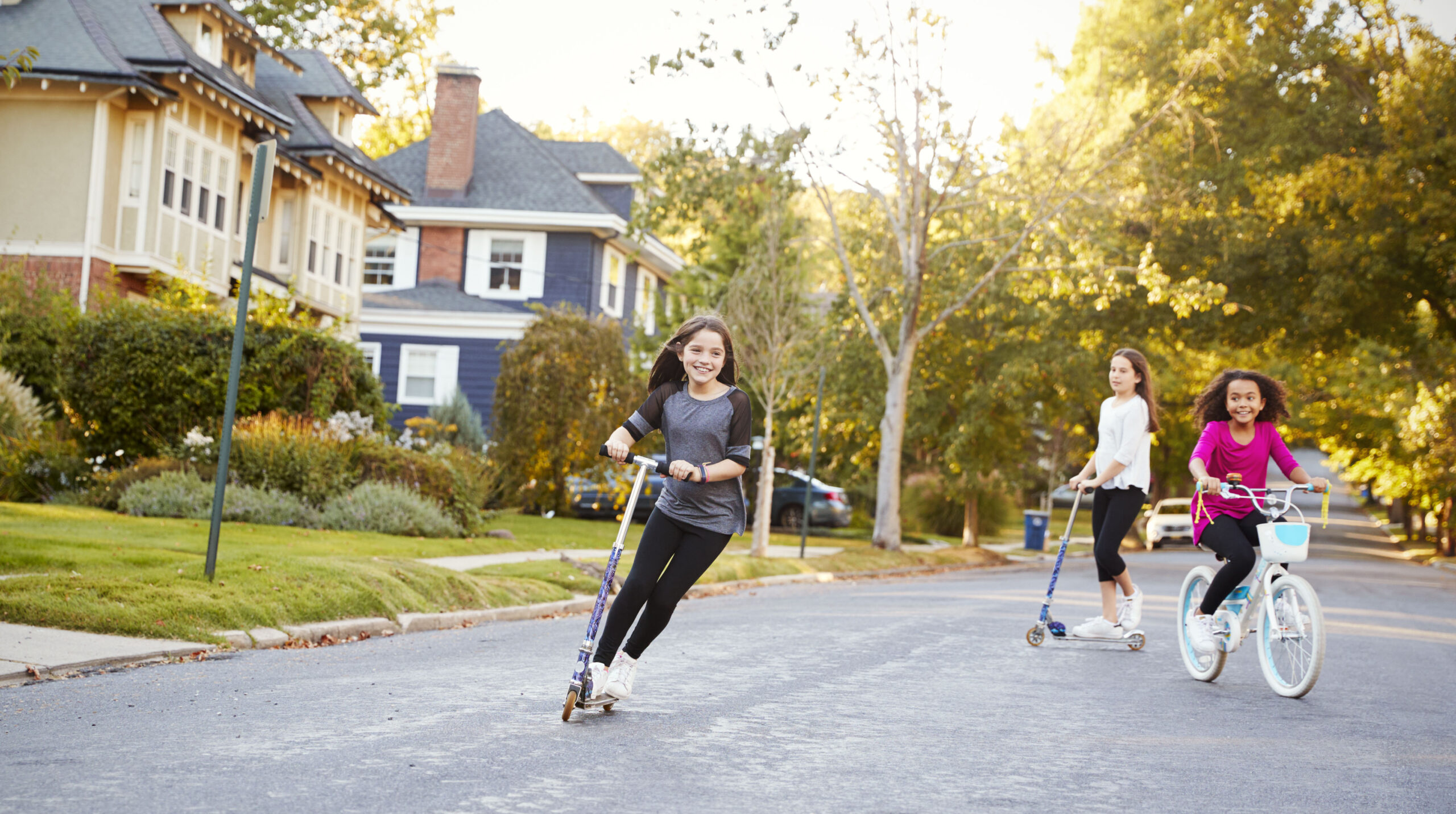 Three pre-teen girls playing in street on scooters and bike