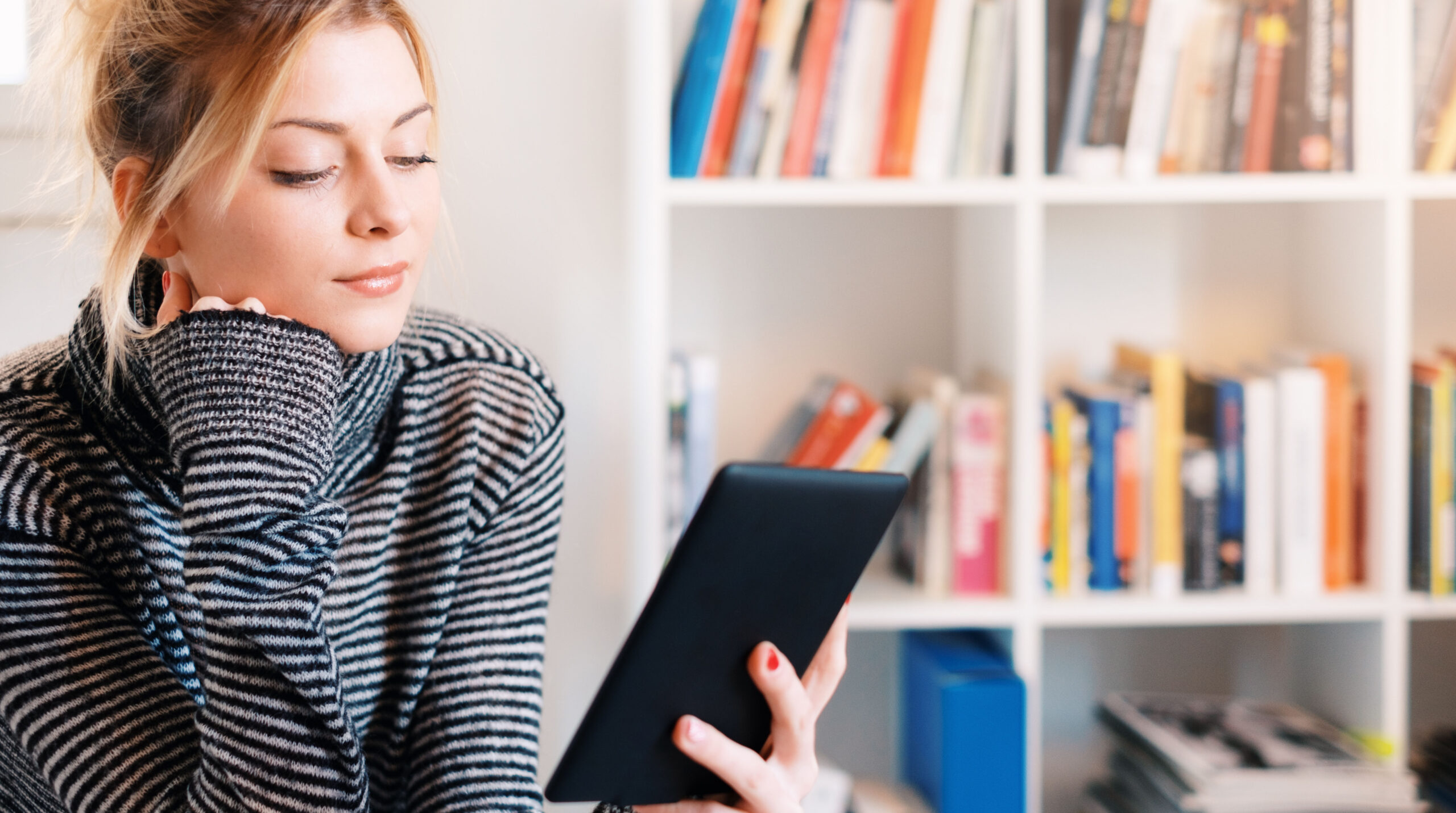 Young girl reading e-book reader next to colorful bookshelf
