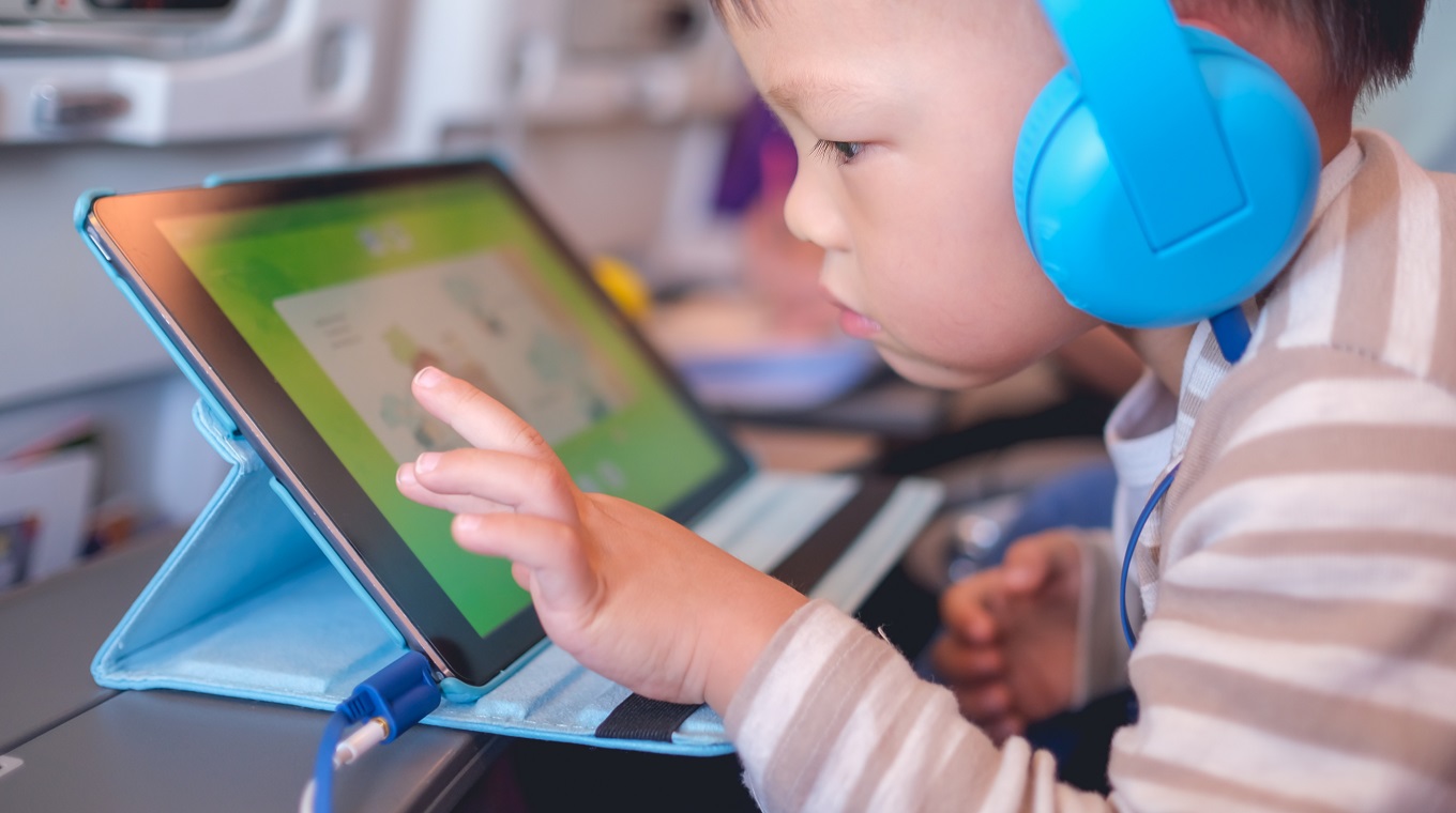 Asian 2 - 3 years old toddler boy child wearing headphones using tablet pc watching cartoons / playing game during flight on airplane