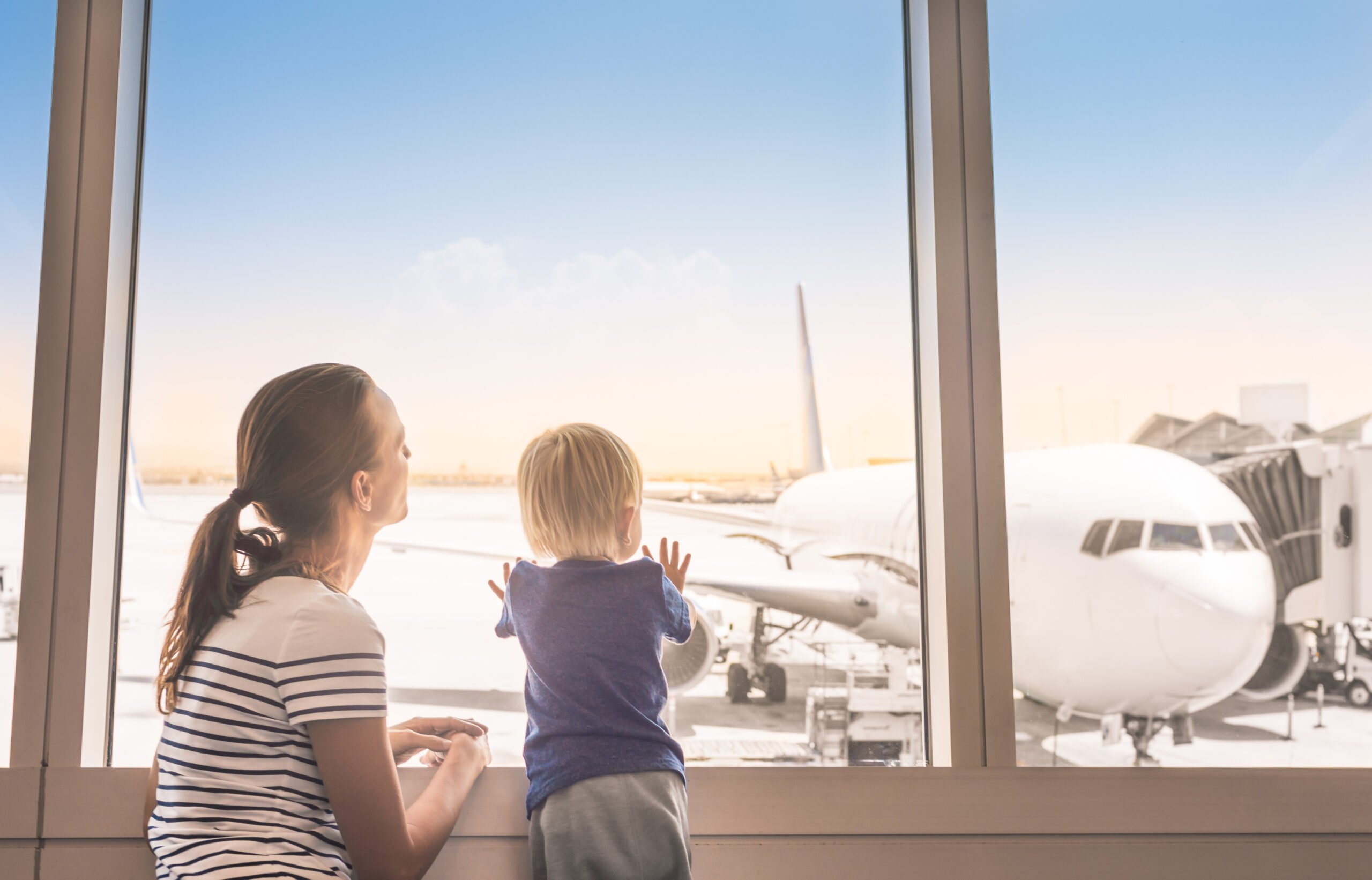 Mother and son at airport boarding terminal looking out window at airplane. Family travel and adventure.