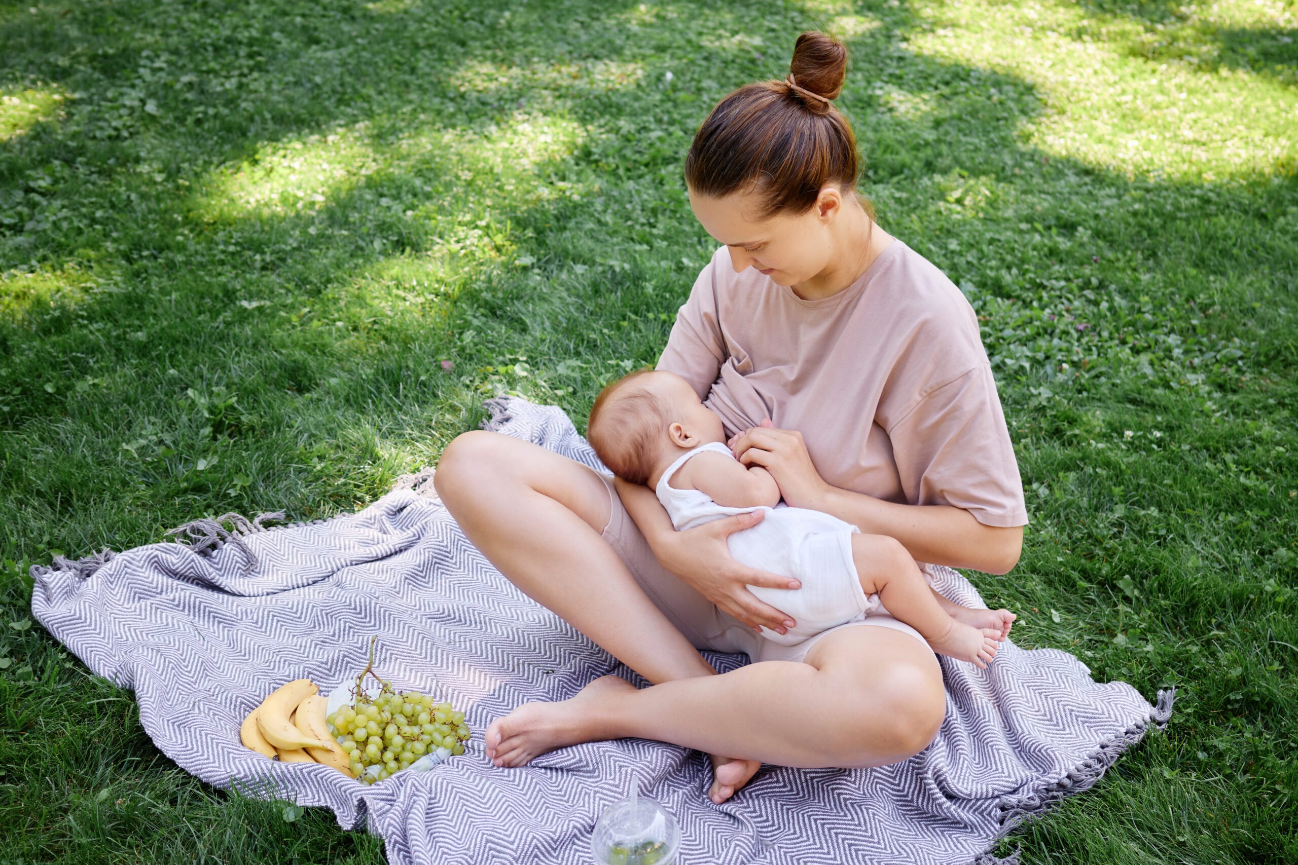 Cheerful mother breastfeeding her newborn son during family picnic in the park sitting on blanket surrounded by green grass on summer day with her child looking satisfied