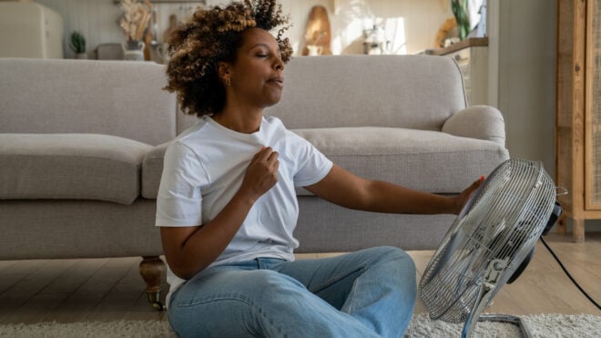 Young African American woman cooling down by ventilator at home, feeling unwell during heatwave