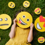 The child covers his face with a emoticon showing his tongue. The person is lying on the grass with emoticons with different psychological moods.