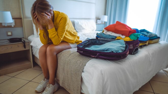 Flight cancellation. Upset woman sitting on bed with suitcases with clothes.
