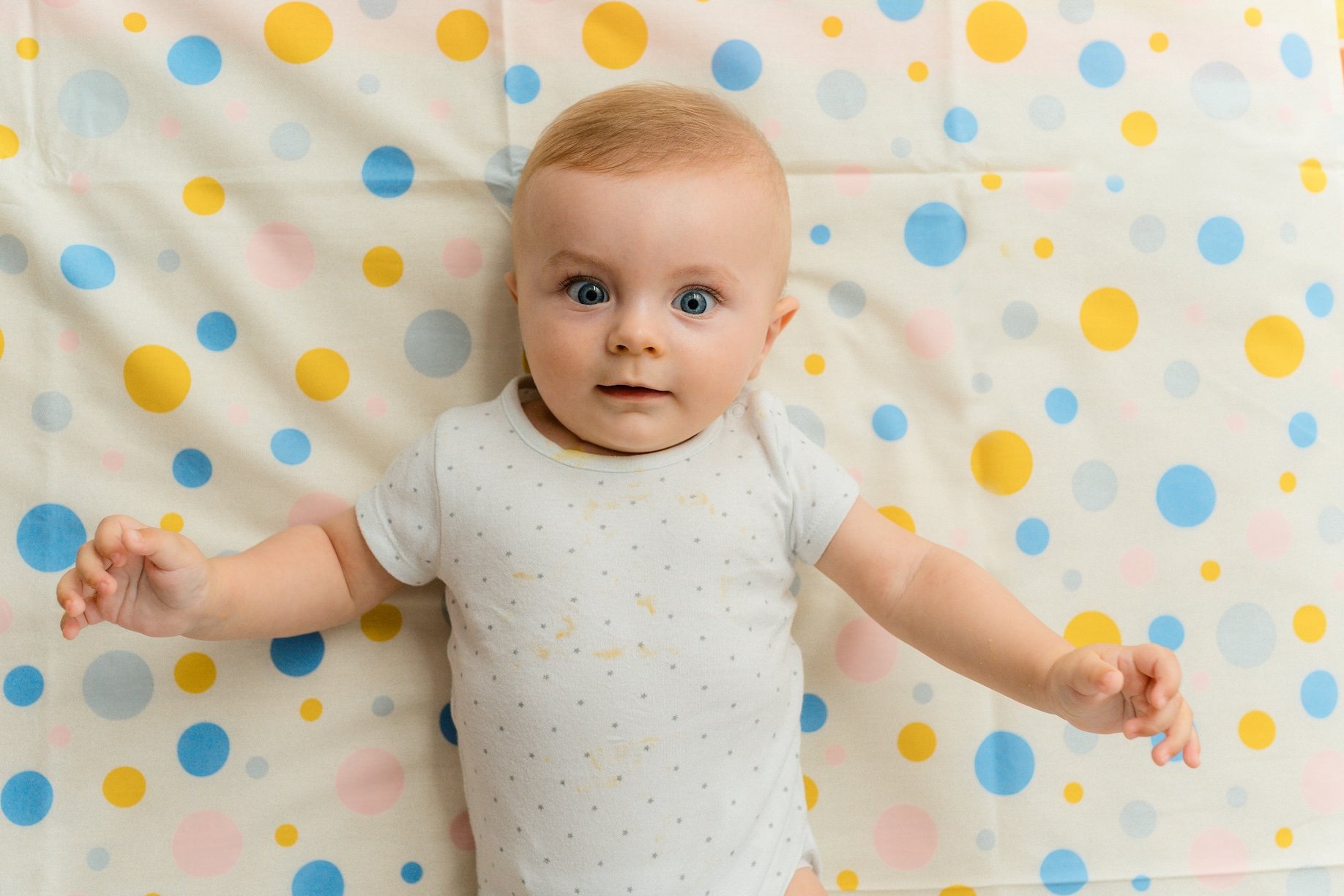 White baby wide-eyed laying on colorful sheet.