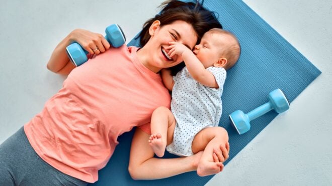 A smiling young mom laying on a blue fitness mat with a baby with small weights beside them