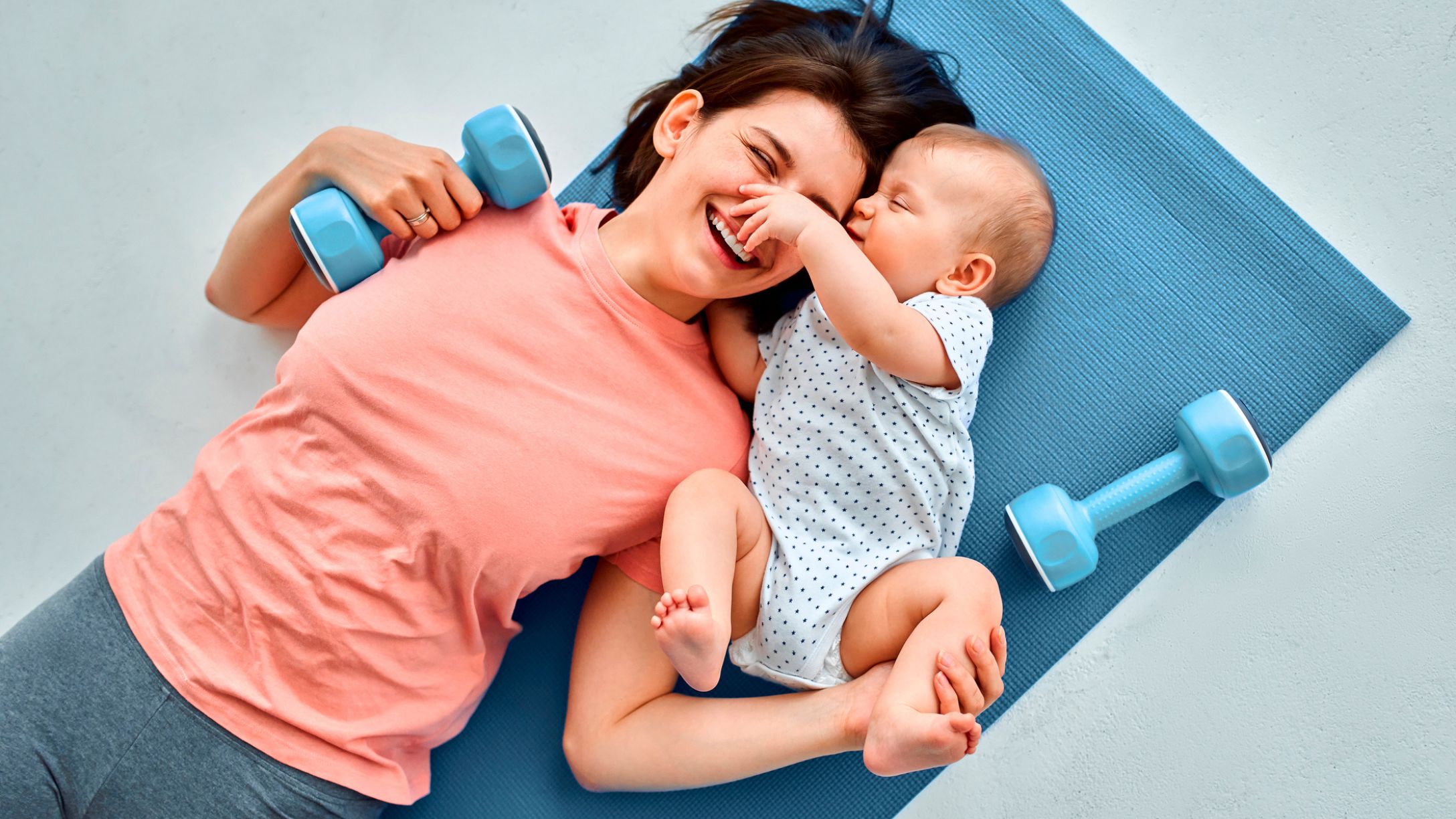 A smiling young mom laying on a blue fitness mat with a baby with small weights beside them