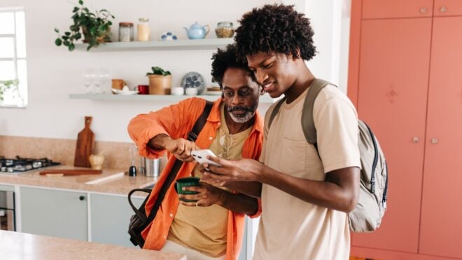 Dad and teen son on the phone together in their kitchen.