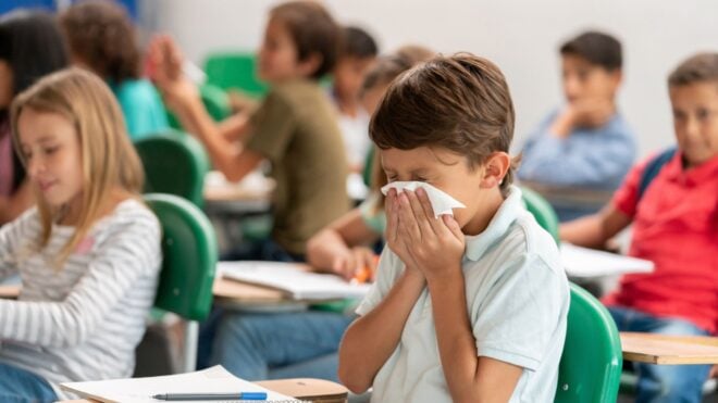 A boy blowing his nose at his desk in a classroom full of kids