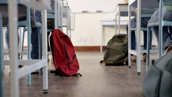 A photo of students’ desks and feet and their backpacks on the floor in a classroom