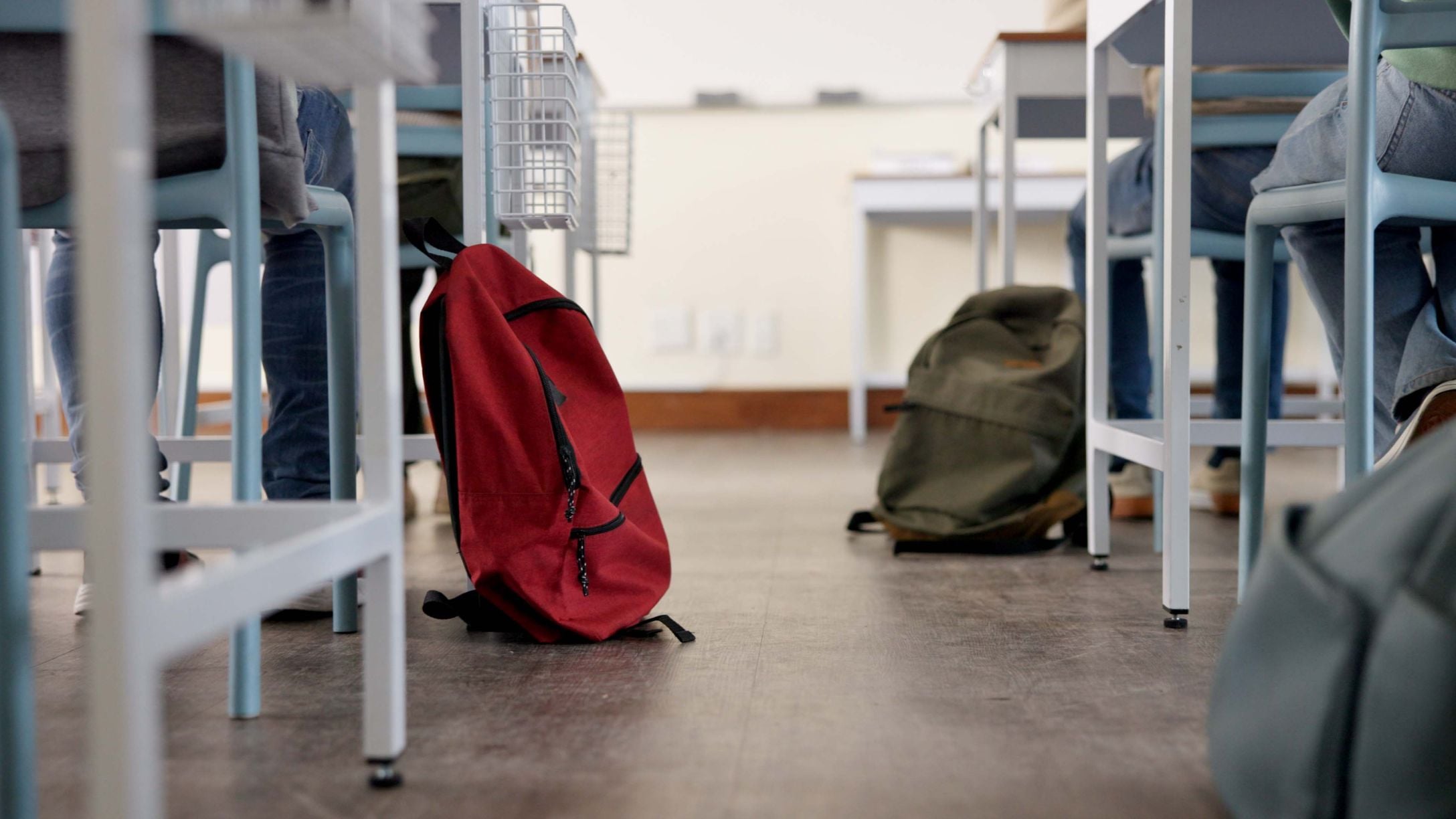 A photo of students’ desks and feet and their backpacks on the floor in a classroom