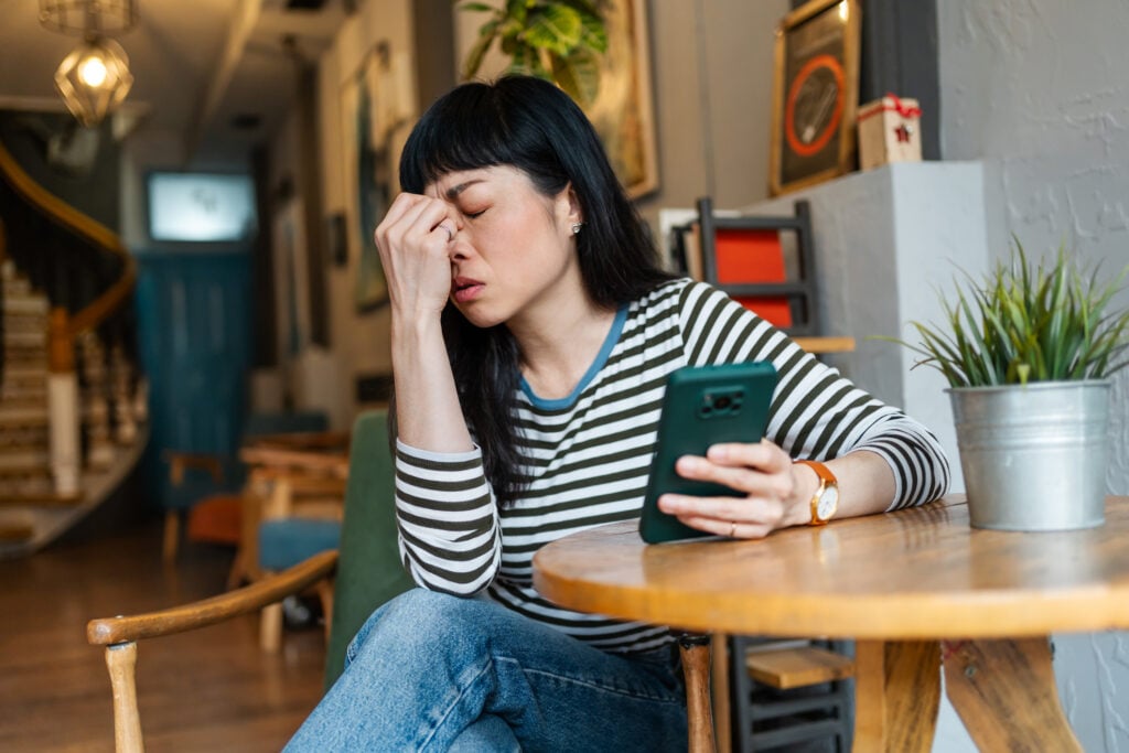 Young Asian woman is trying to contemplate lousy news she is reading online on her smartphone while sitting in a caf&eacute;