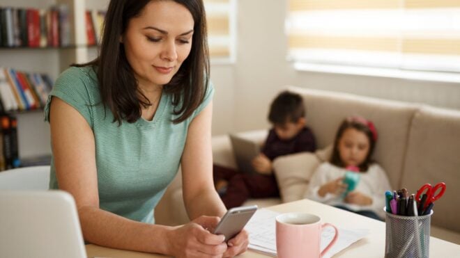 A mother on her phone and laptop working from home with her kids on the sofa behind her