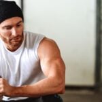 A photo of a man in a sleeveless top with a hat on showing off his muscles