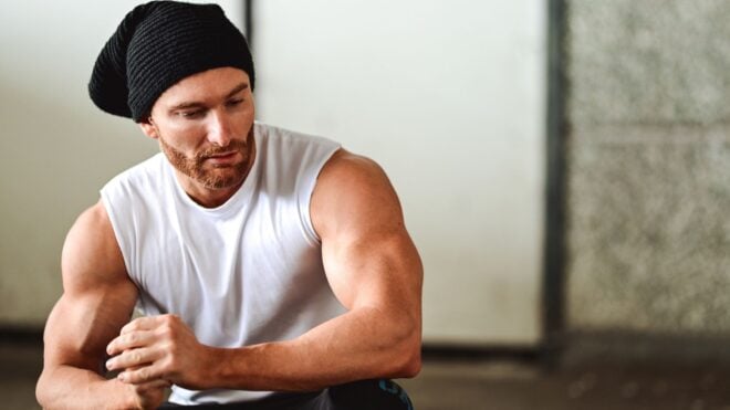 A photo of a man in a sleeveless top with a hat on showing off his muscles